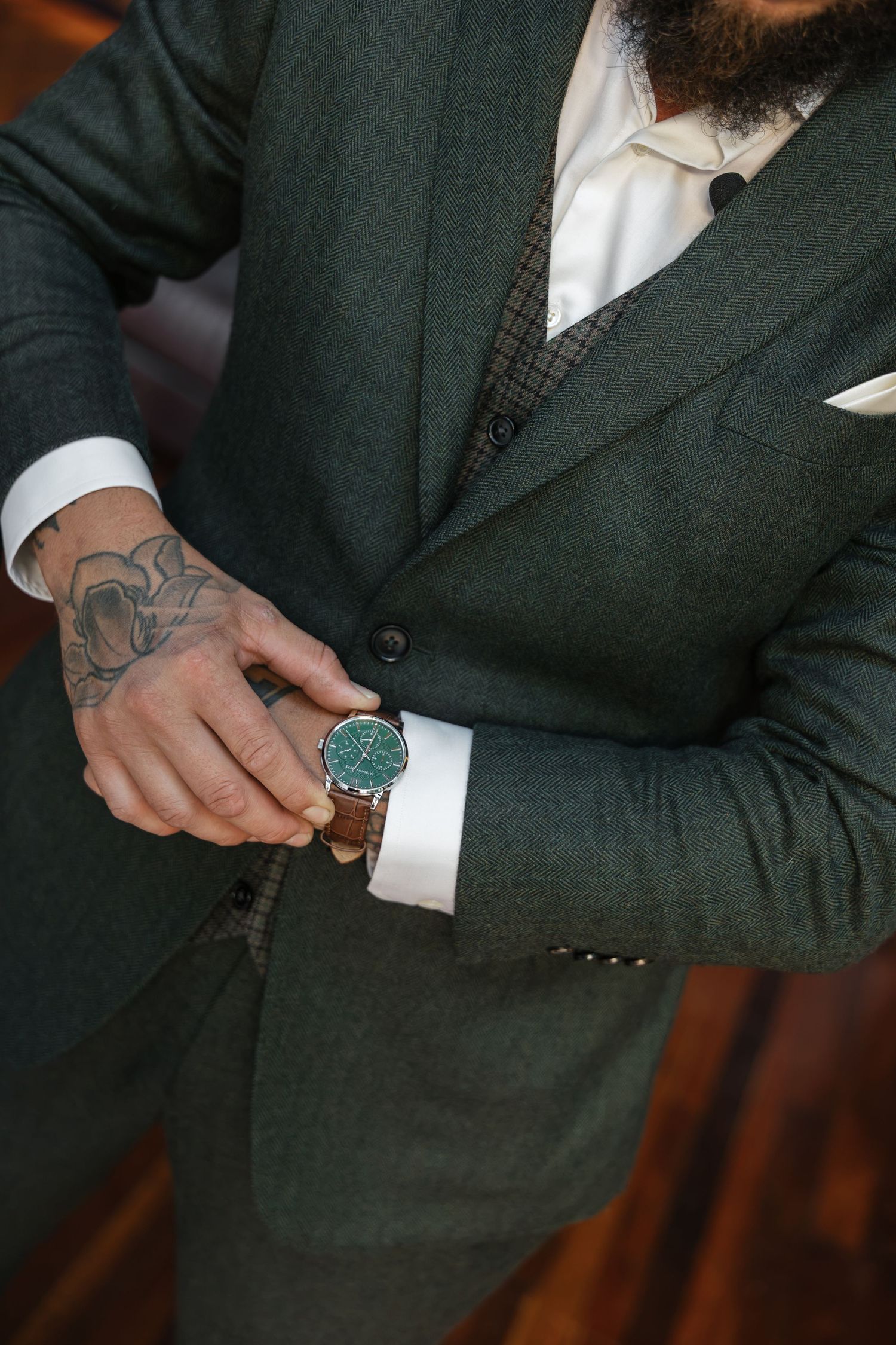 Man in green suit adjusting brown leather watch with green dial, tattooed hand visible, white dress shirt cuff showing.