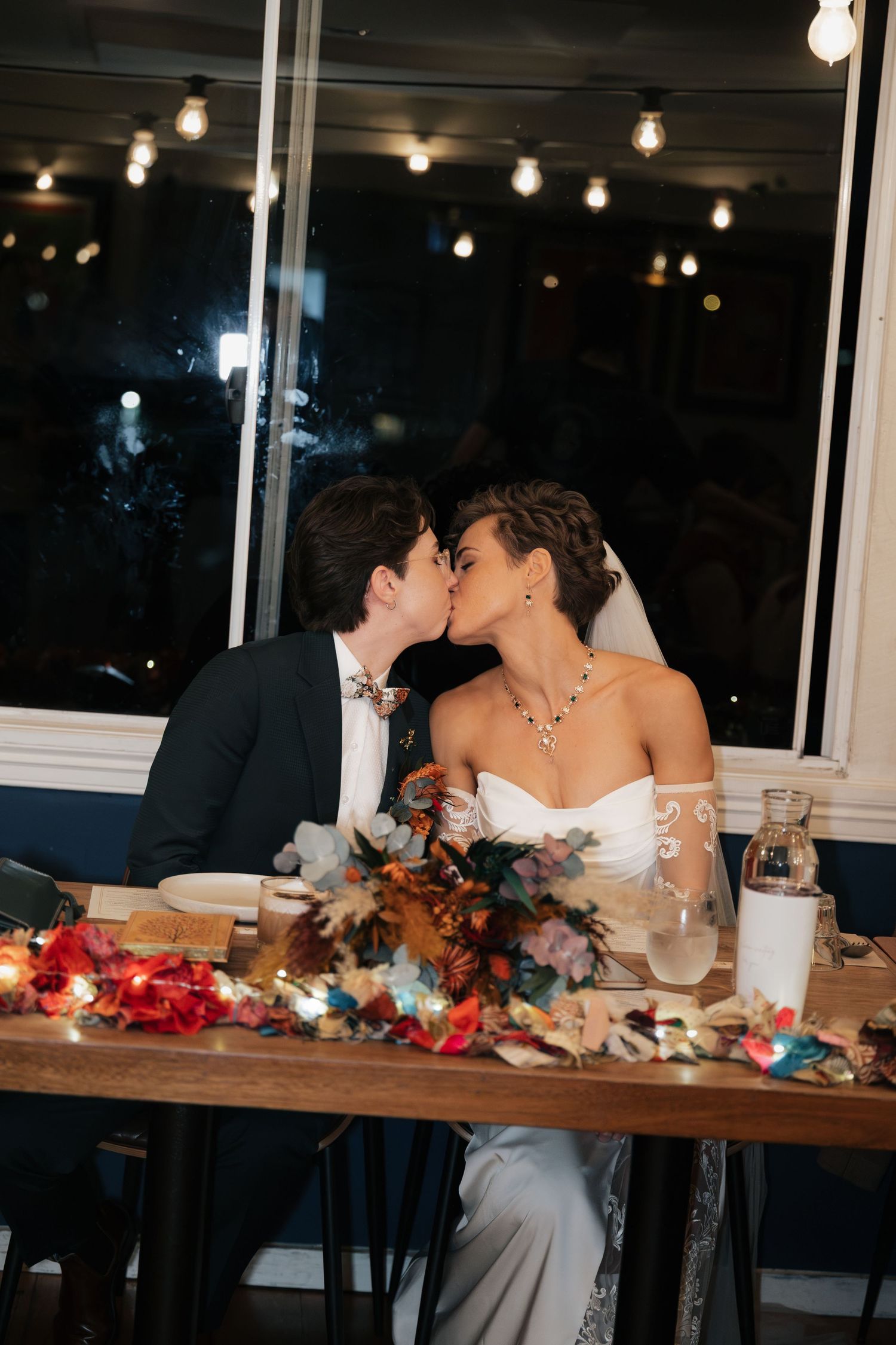 Newlywed couple sharing a kiss at their wedding reception dinner table adorned with colorful floral arrangements and string lights.