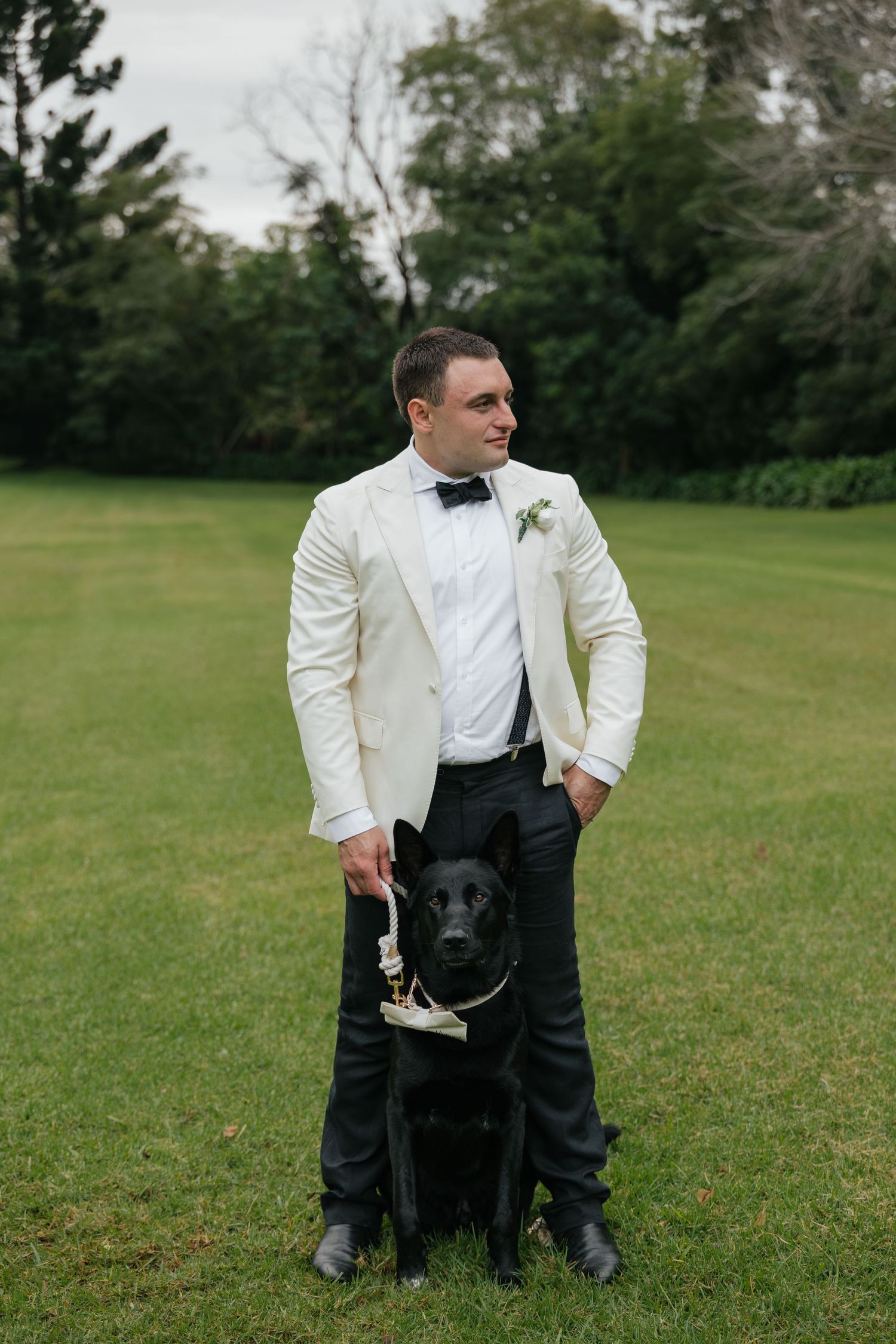 Groom in white tuxedo jacket and bow tie stands with black Labrador on green lawn with trees behind.