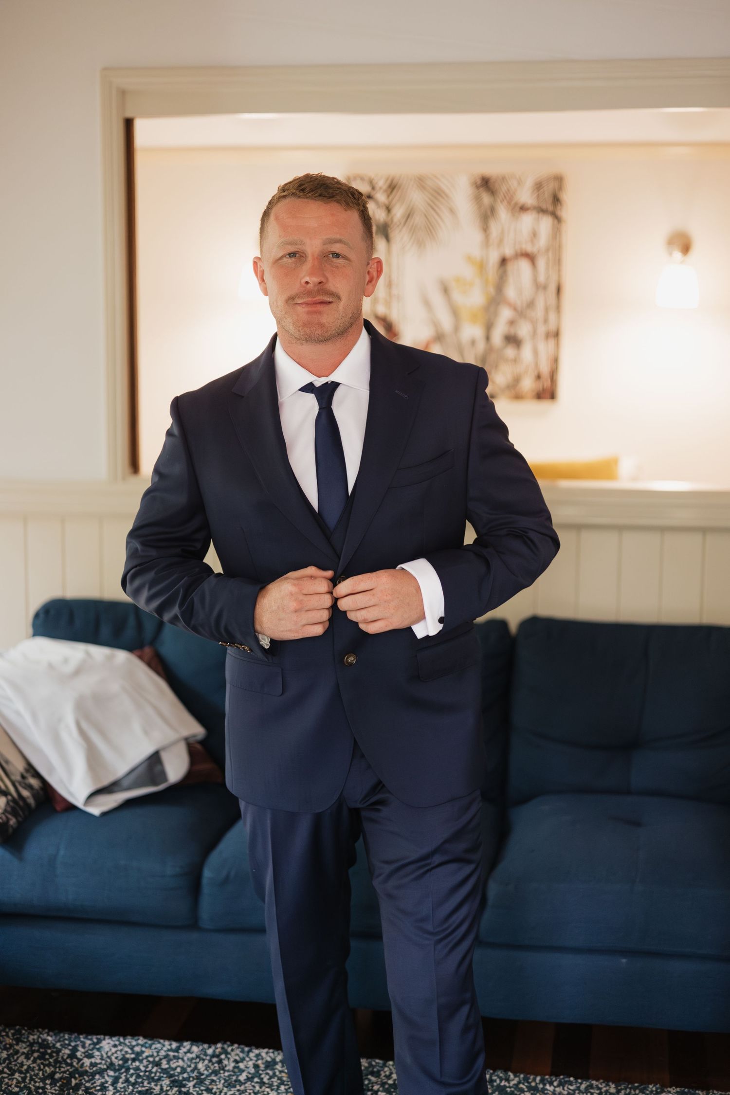 A man in a navy blue suit and tie buttoning his jacket while standing in an elegantly furnished room.