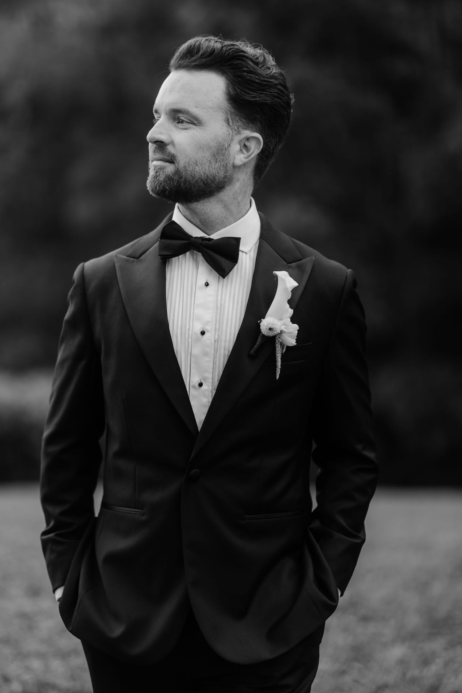 Black and white portrait of a bearded groom in a tuxedo with bow tie and boutonniere, hands in pockets outdoors.