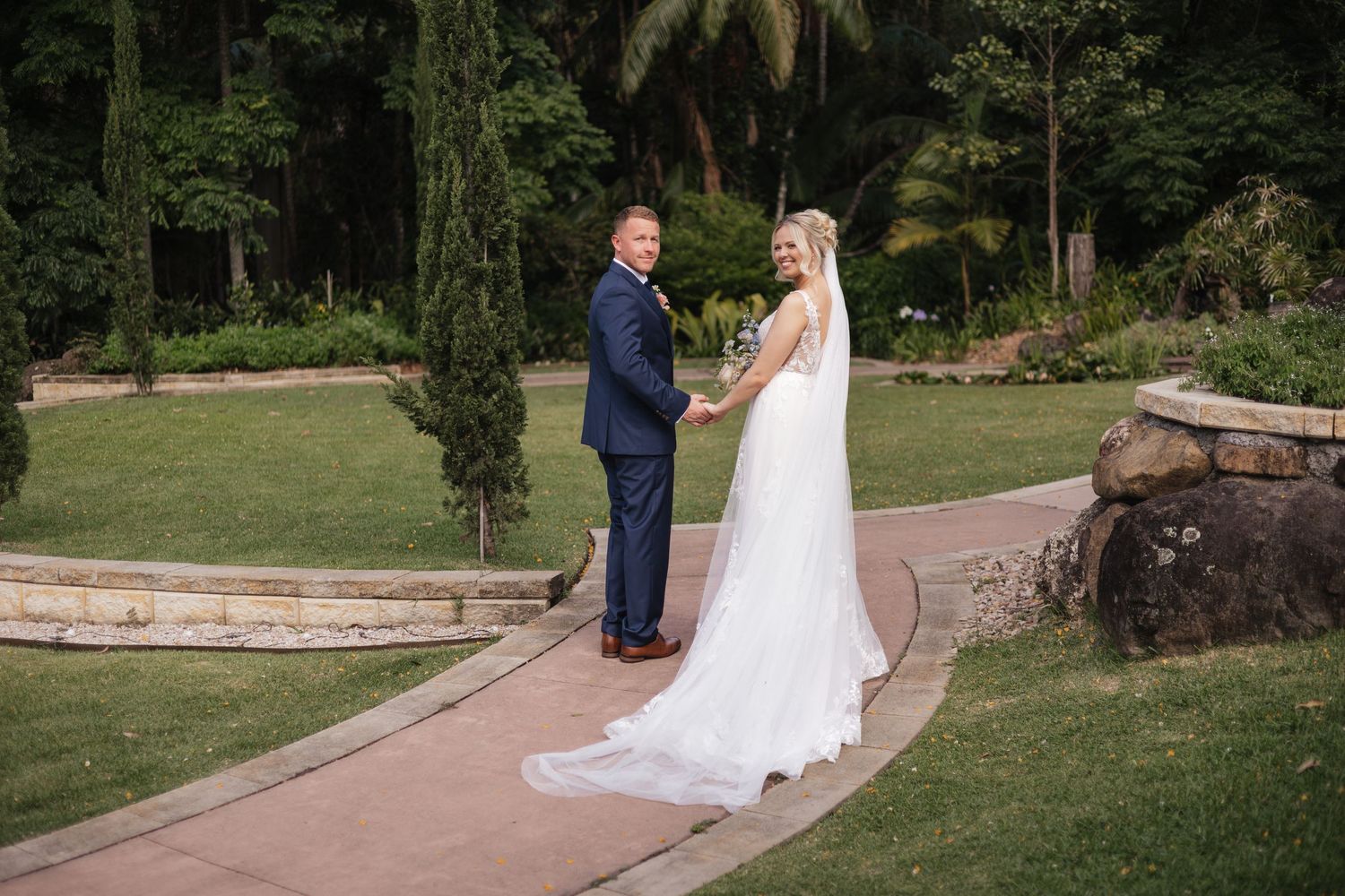 Bride in white gown and groom in navy suit hold hands on garden path surrounded by lush greenery on their wedding day.