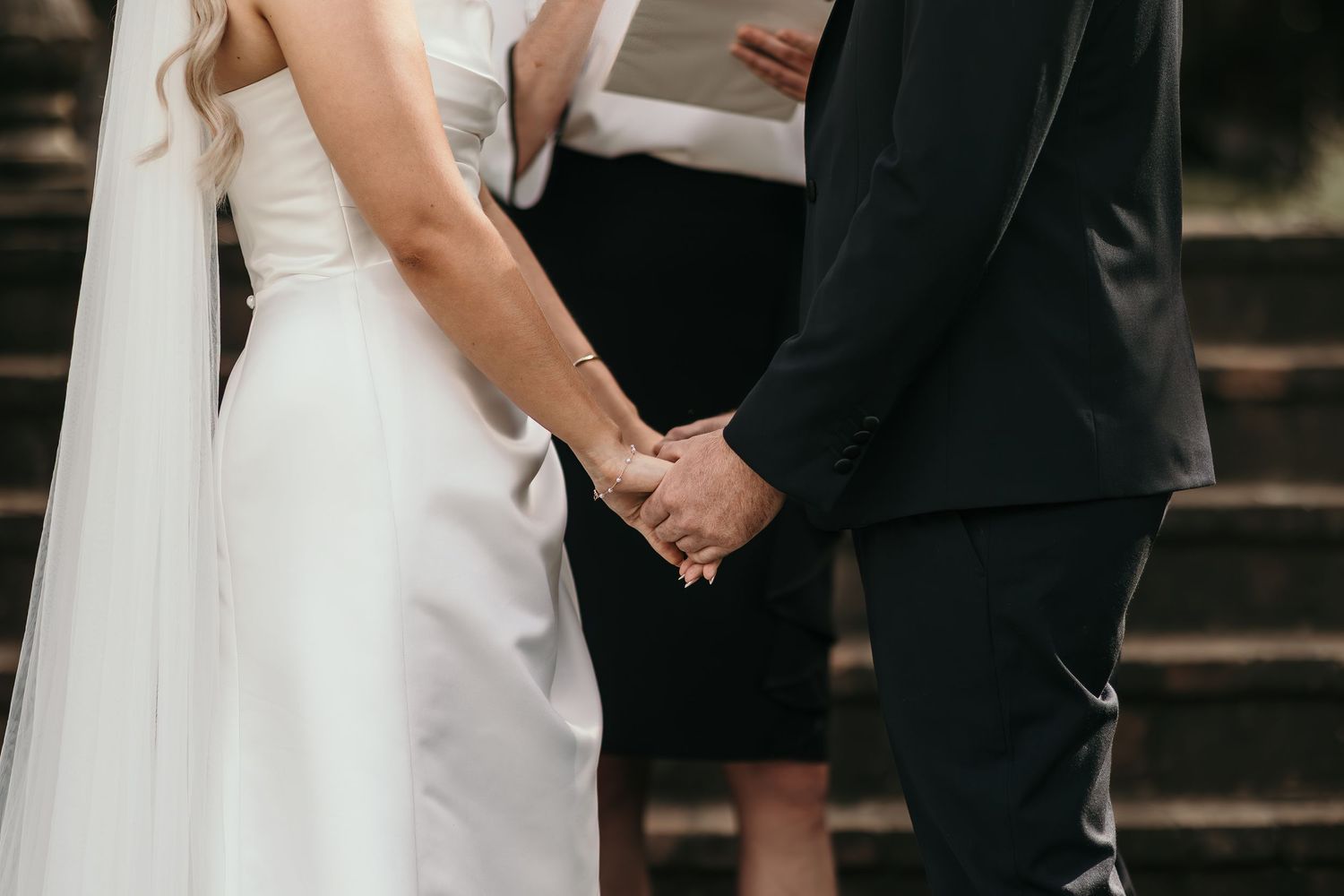 Bride and groom holding hands during wedding ceremony, officiant in background on stone steps.