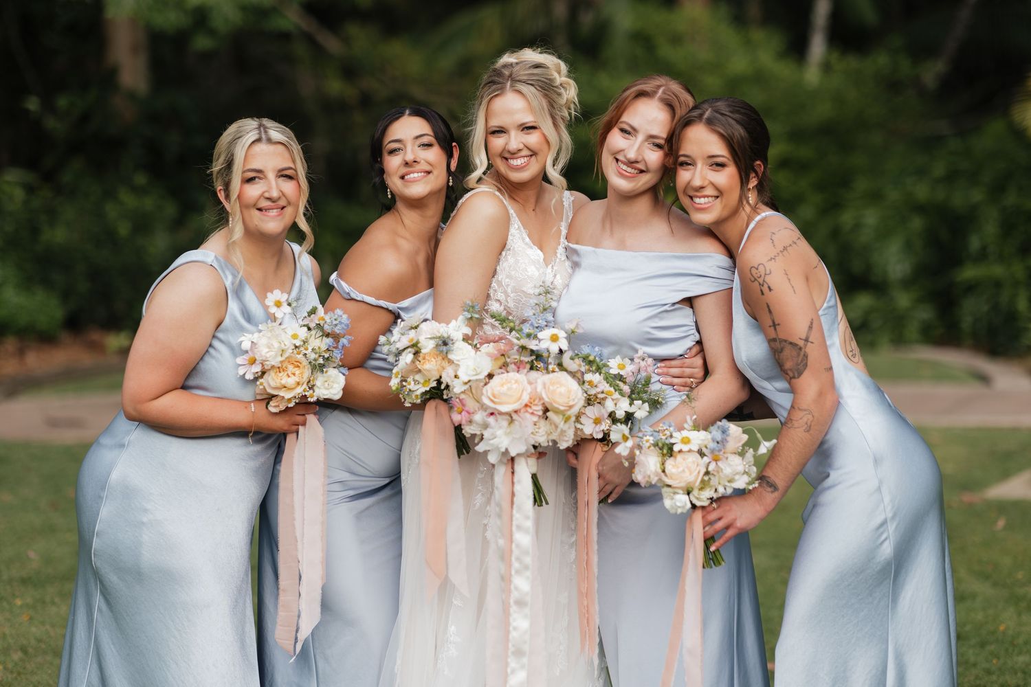 Bride in lace gown poses with four bridesmaids in light blue satin dresses holding pastel bouquets outdoors.