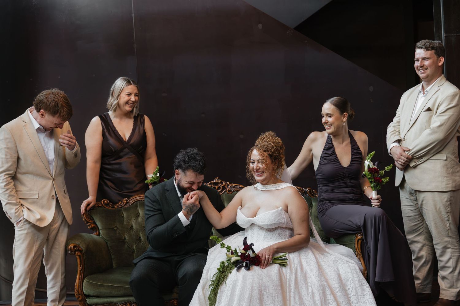 Groom kisses bride's hand while wedding party laughs and celebrates together in a stylish dark-themed venue.