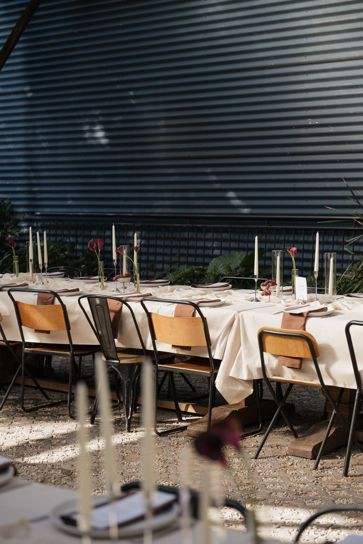 Elegant outdoor dining table set with white linens, candles, and floral accents on gravel patio.