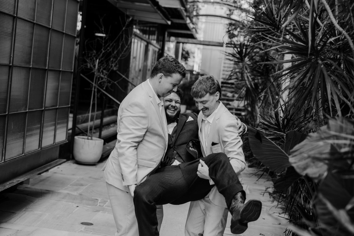Three groomsmen laughing and joking together outdoors in black and white wedding photo.