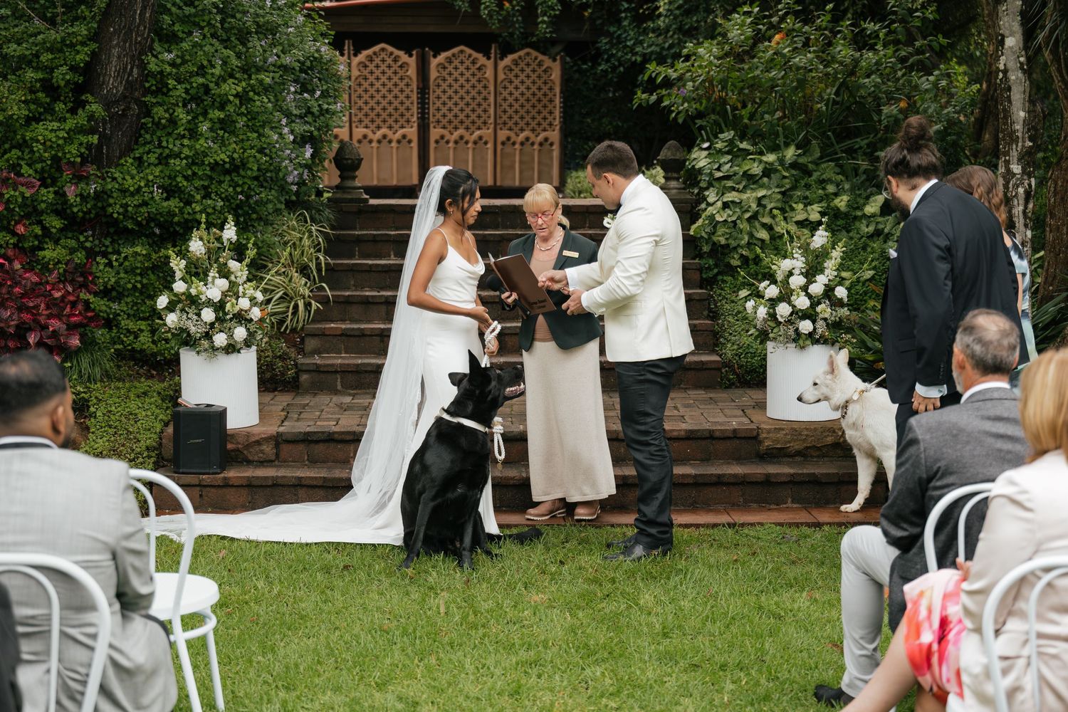 Outdoor wedding ceremony with bride, groom, officiant, and black dog on green lawn with floral arrangements and seated guests.