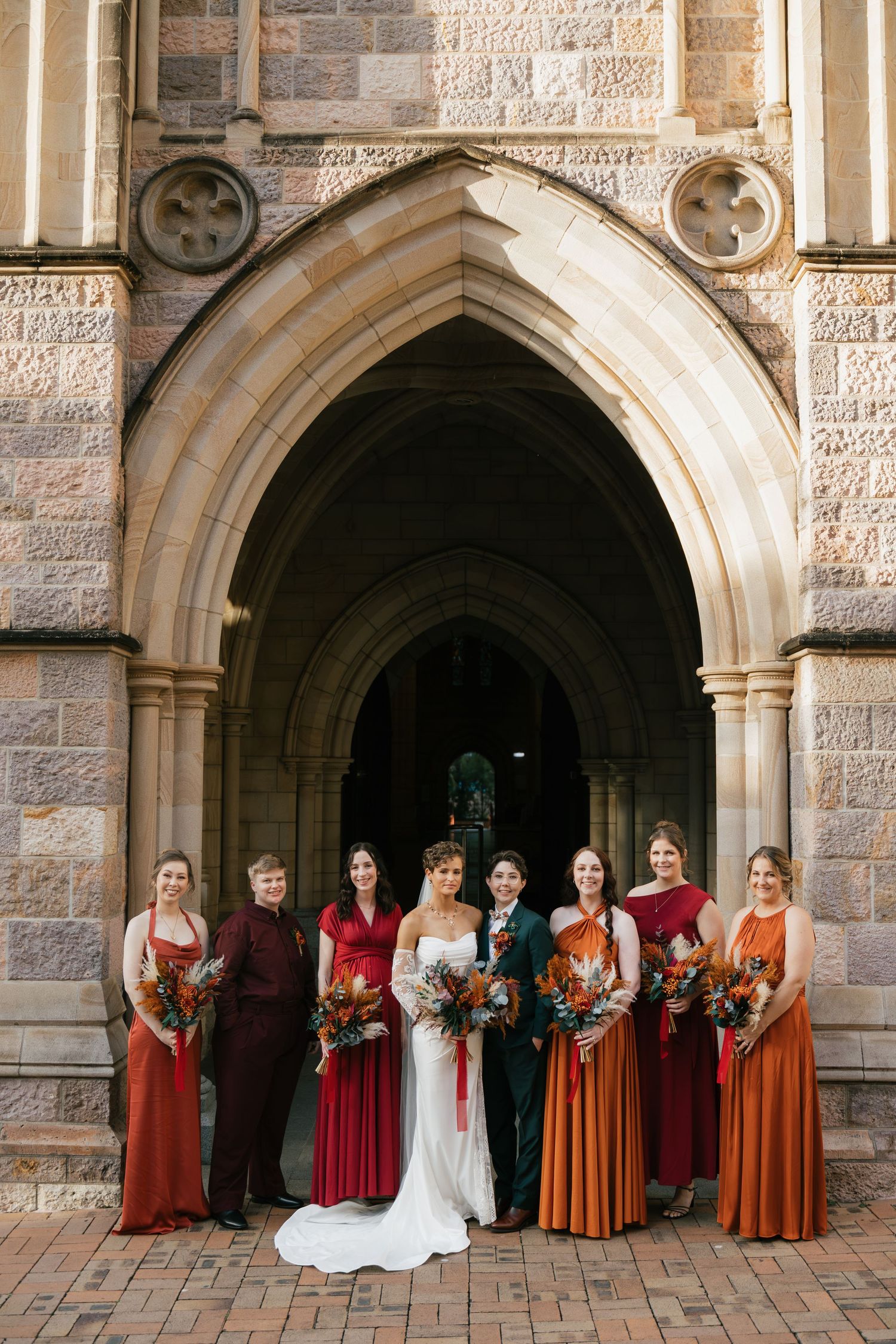 Wedding party poses in autumn-toned dresses before a Gothic stone church arch, with bride in white gown.