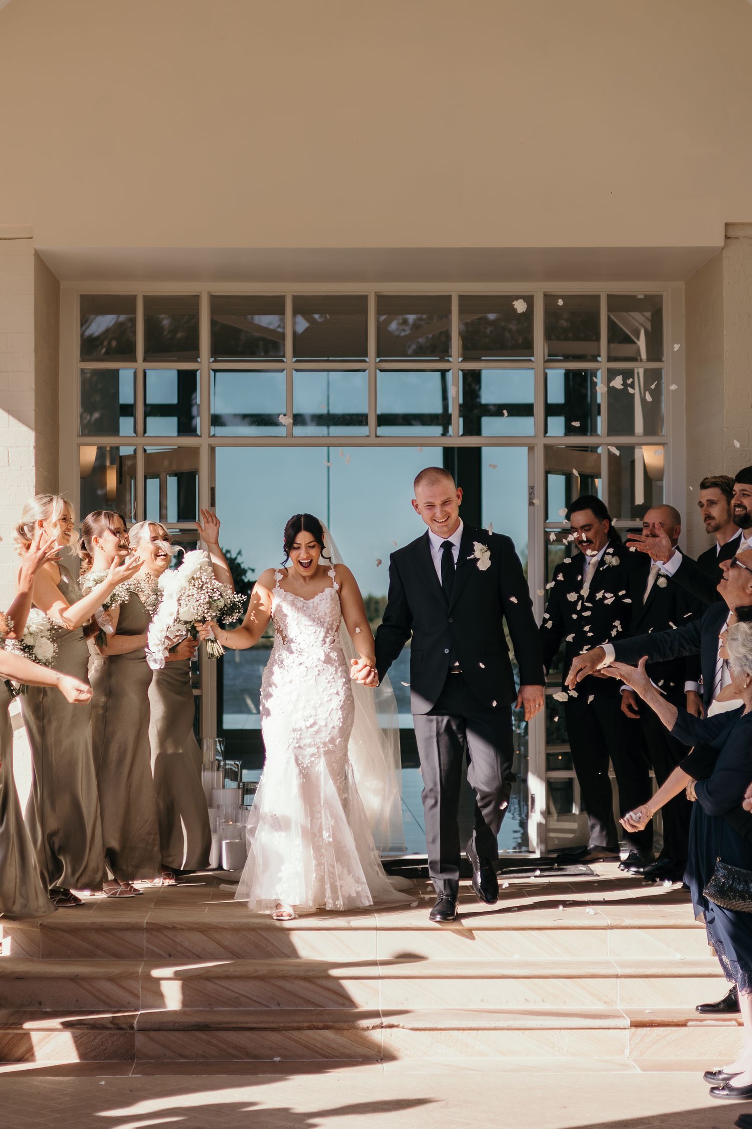 Wedding guests line hallway as newlyweds exit ceremony venue while bubbles float in afternoon sunlight.