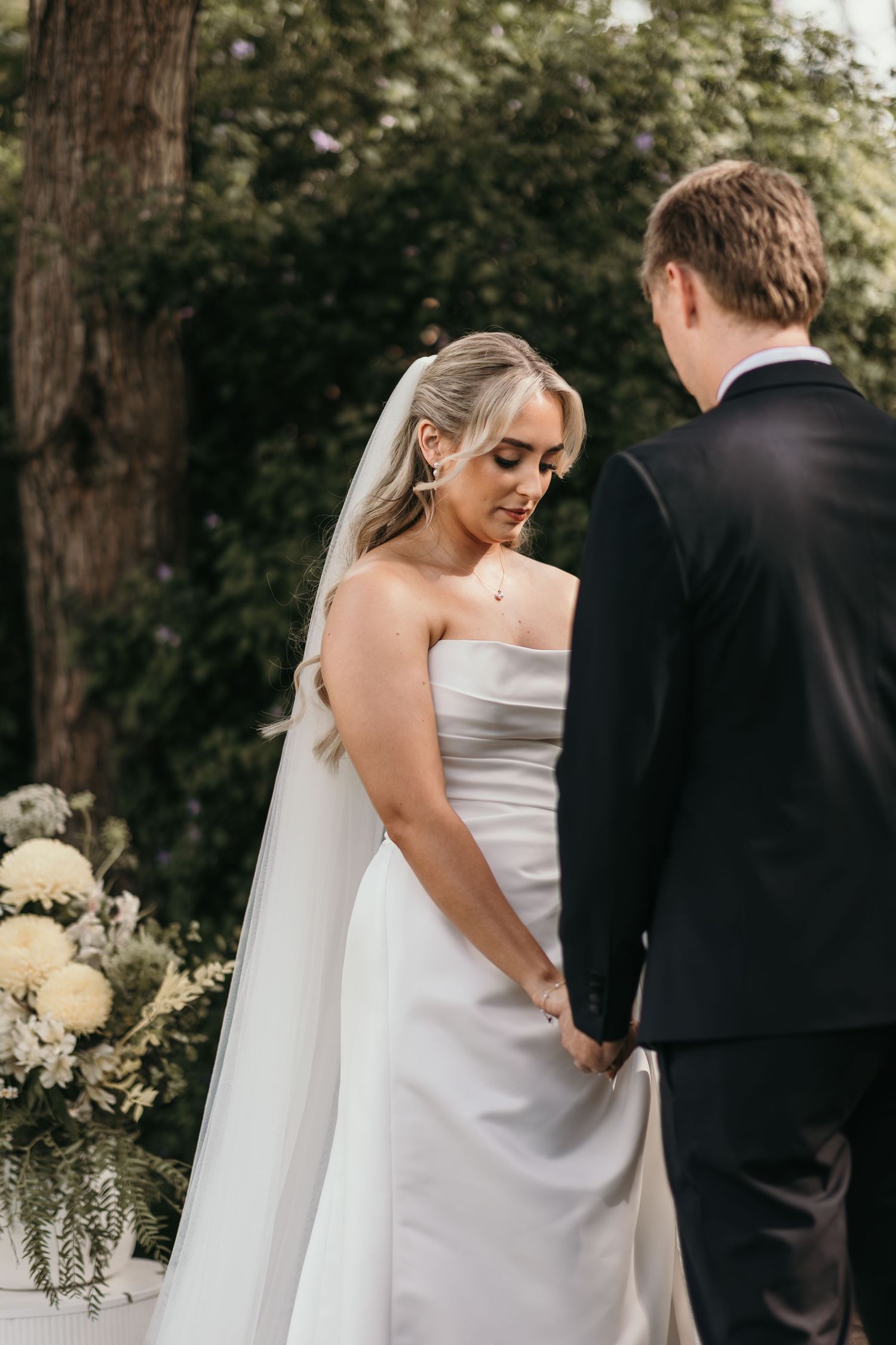 Bride in strapless white gown and veil holds hands with groom in black suit during outdoor wedding ceremony.