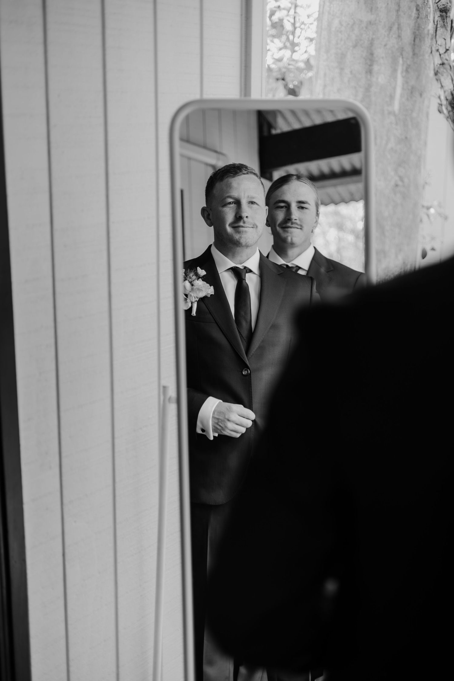 Black and white photo of a groom in a suit with boutonniere reflected in a mirror with his groomsman.