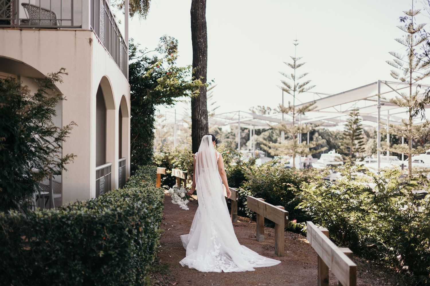 A wedding party walks along a covered outdoor pathway with white columns and lush greenery at a Mediterranean venue.