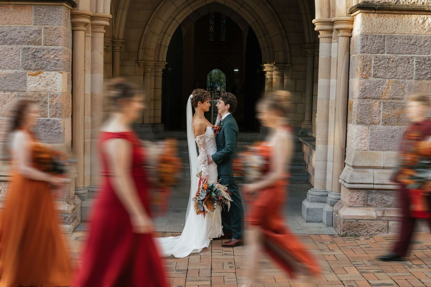 Bride and groom share a romantic moment in a stone archway as bridesmaids in red and orange dresses walk past.