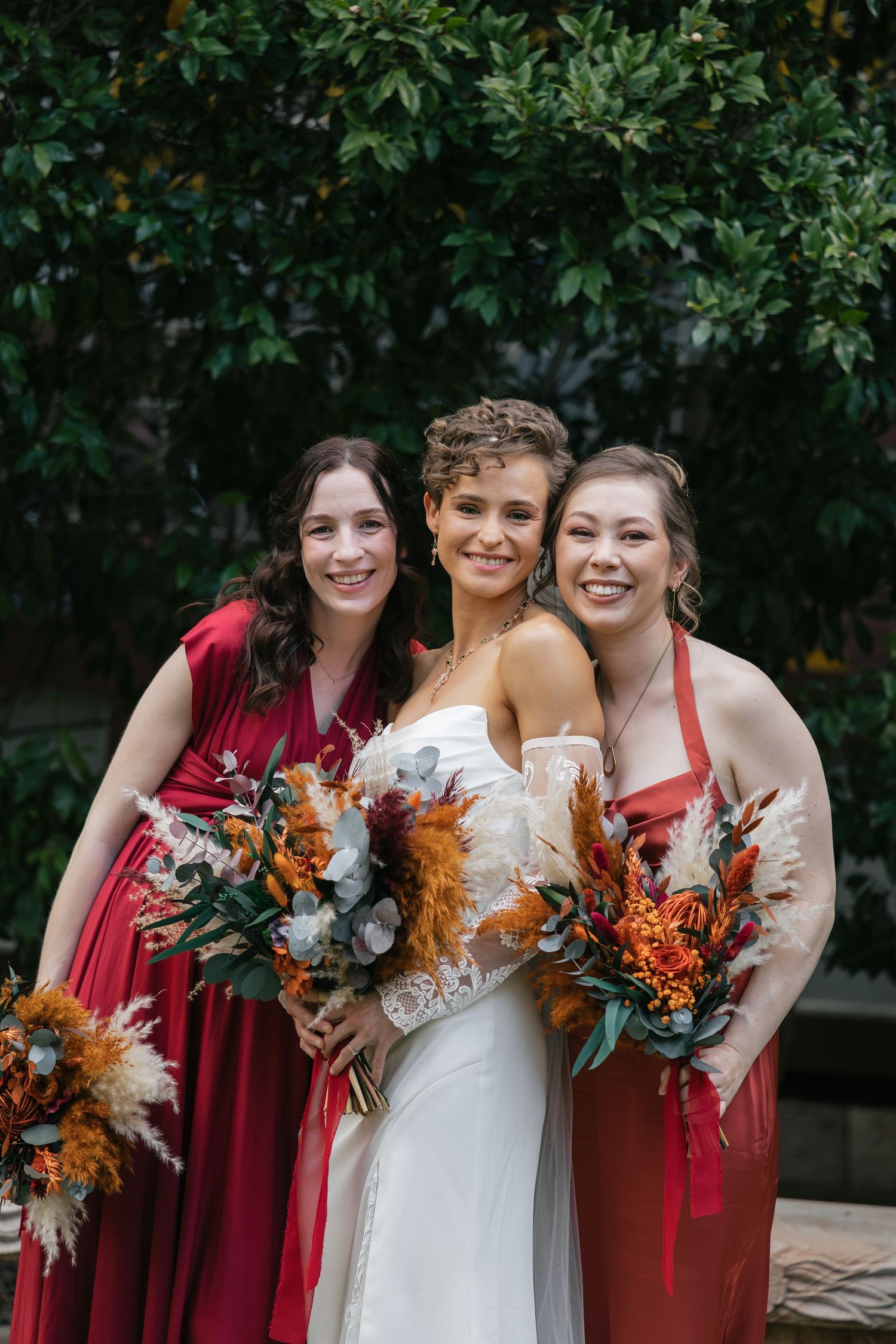 Bride in white gown poses with two bridesmaids in red dresses holding autumn bouquets outdoors.