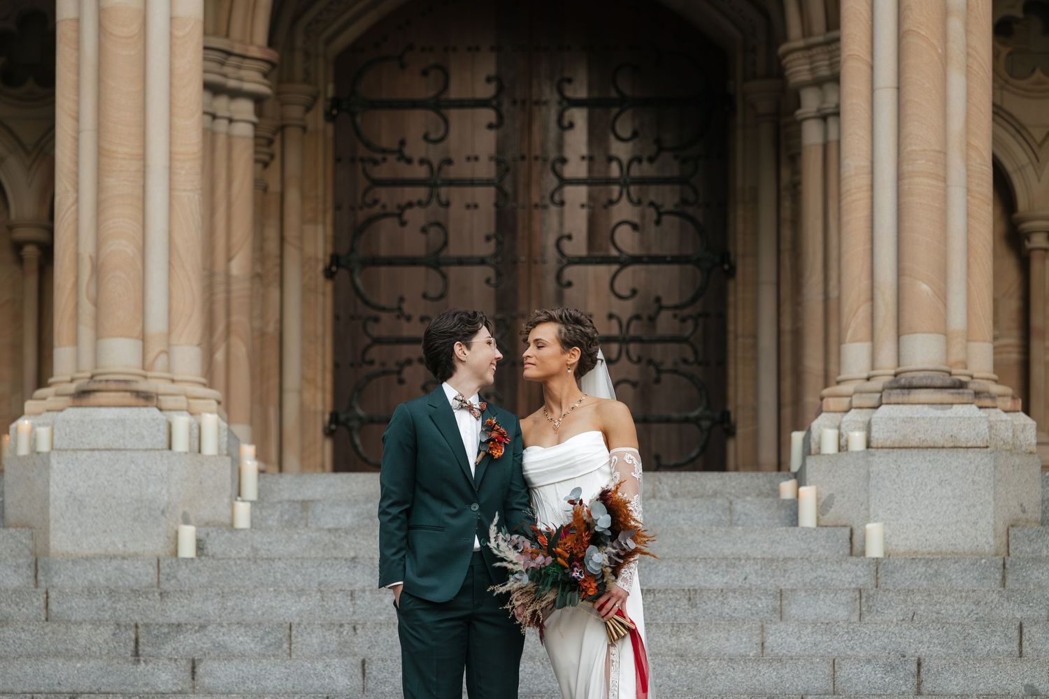 Bride and groom share a romantic moment on church steps, bride holding a fall bouquet with burgundy and terracotta blooms.