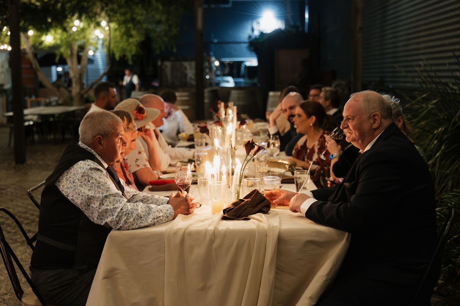 Elegant outdoor candlelit dinner party with guests seated at a long white-tablecloth table at night.