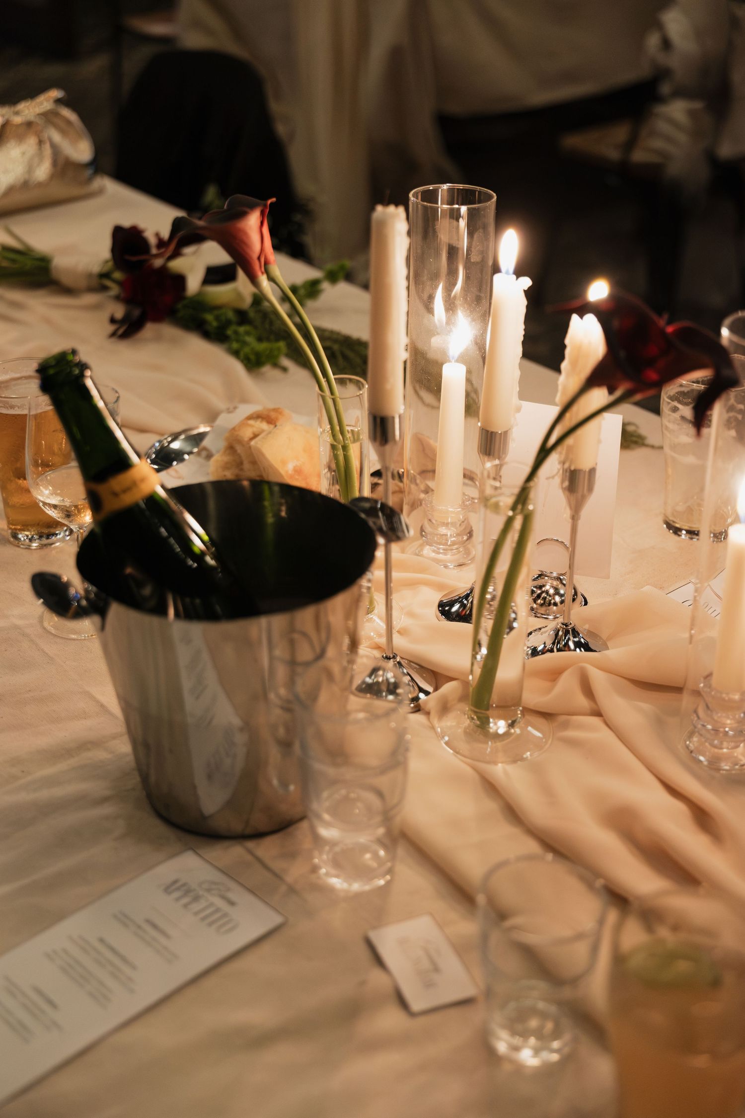 Elegant candlelit dinner table with champagne in ice bucket, white candles, calla lilies, and crystal glassware.