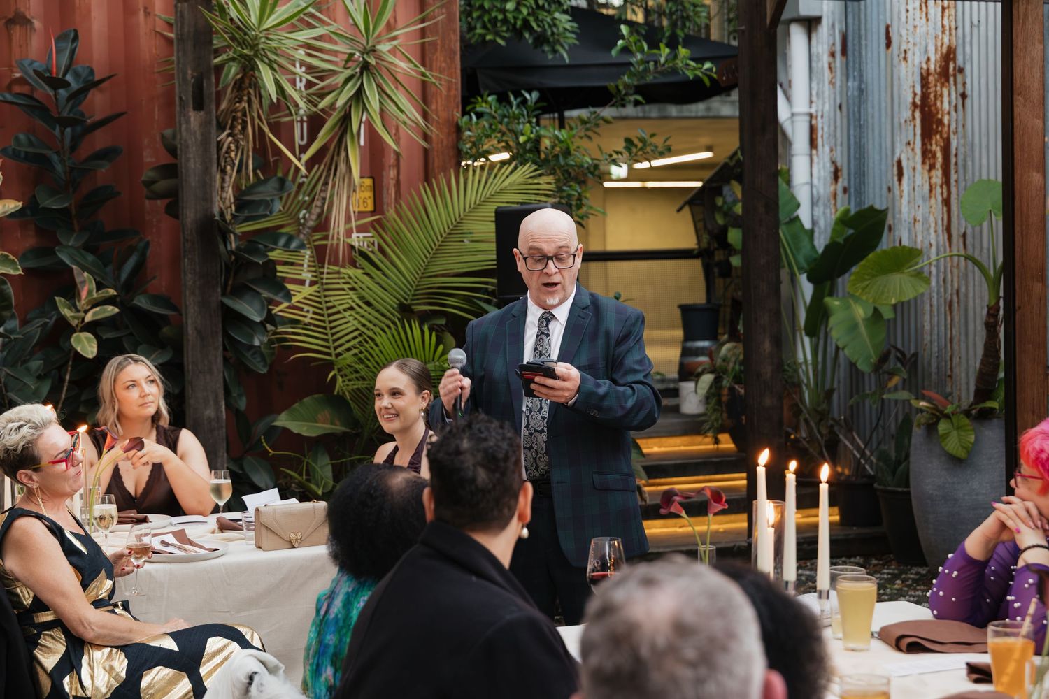 An elderly man in a suit speaks at an outdoor dinner party surrounded by guests and tropical plants.