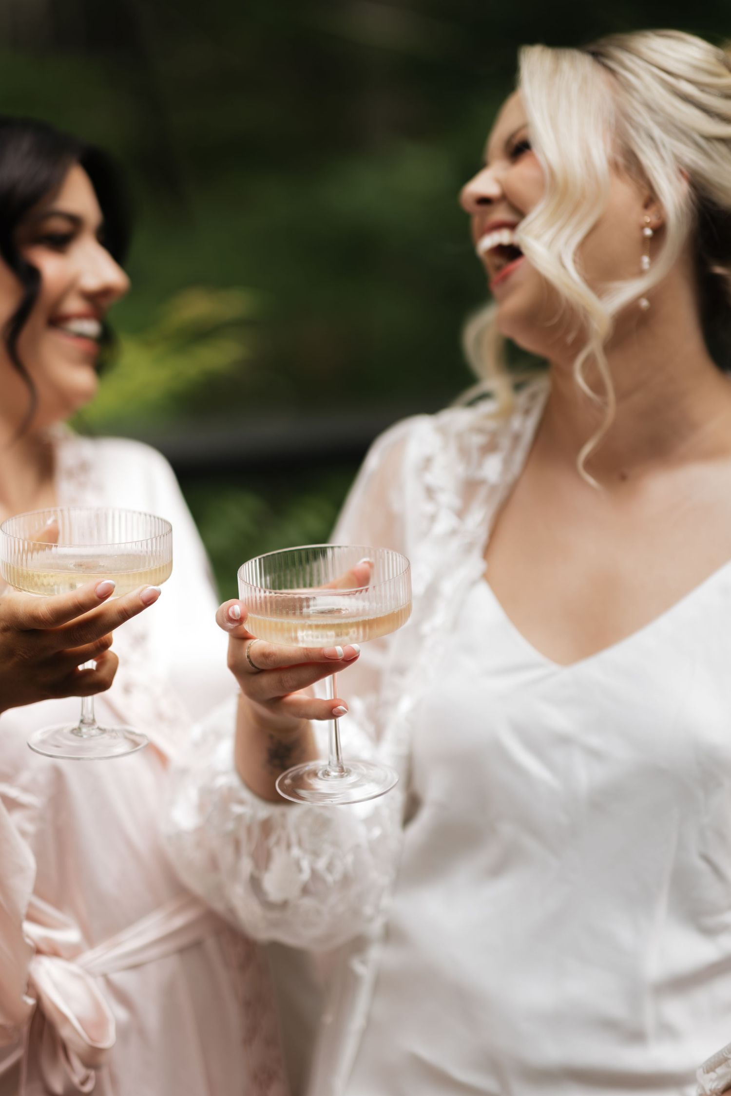 Bride and bridesmaid in white robes laughing and toasting with champagne coupe glasses outdoors.