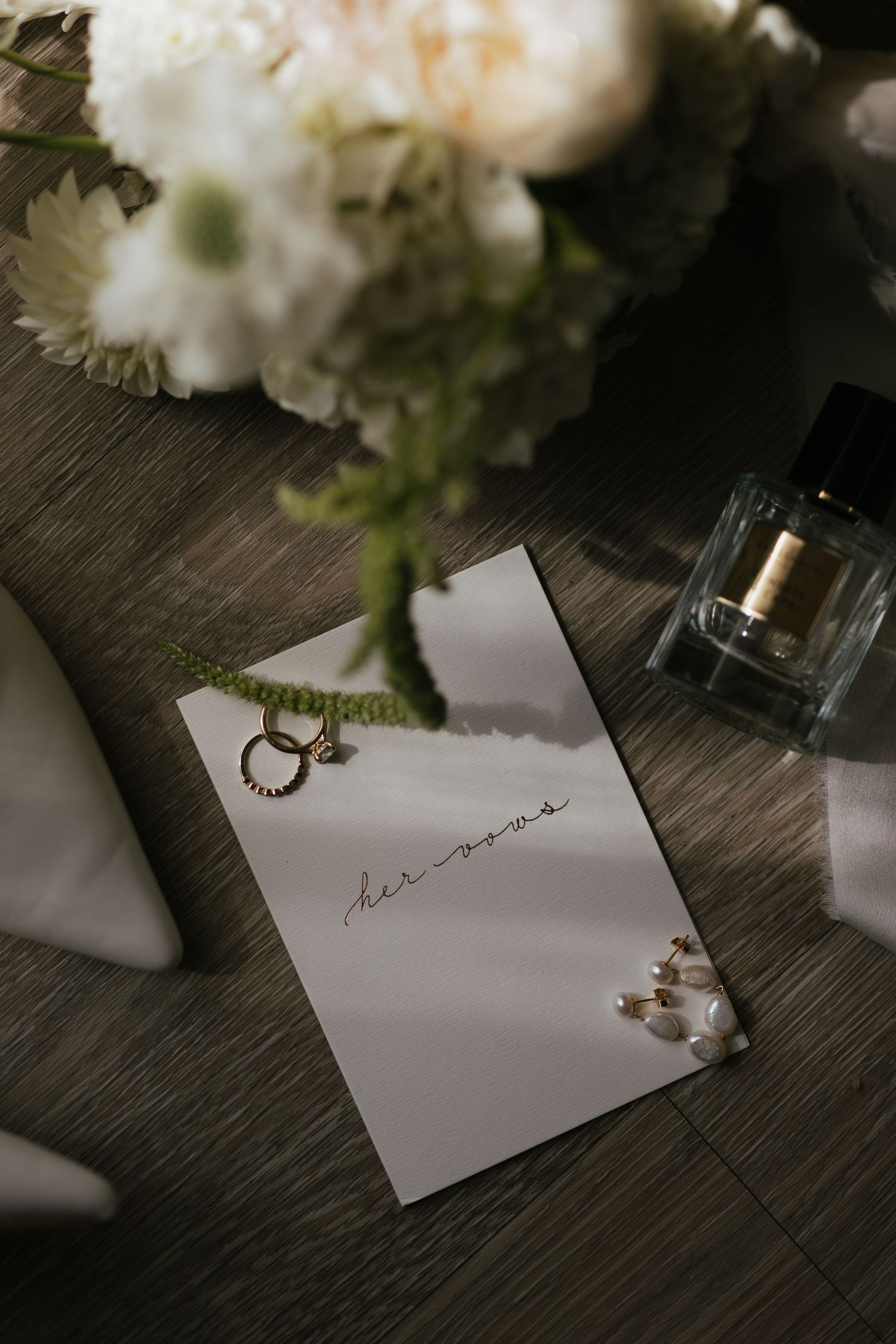 Wedding flat lay with vow card, gold rings, pearl earrings, perfume bottle, and white floral bouquet on wood surface.