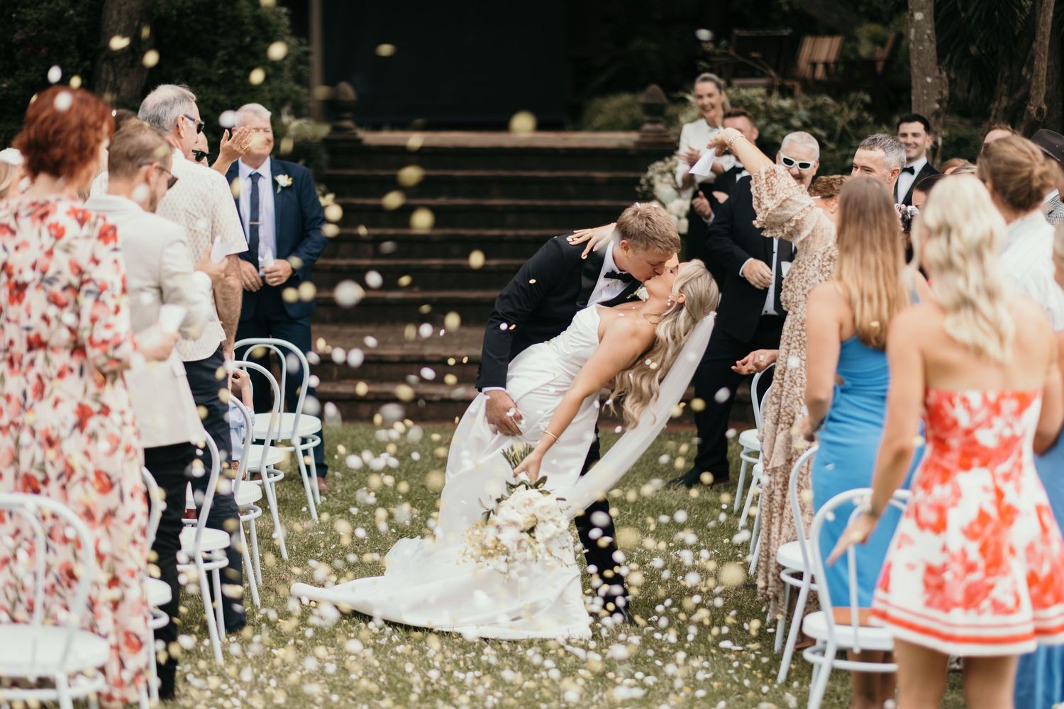 Bride and groom share romantic dip kiss at outdoor wedding ceremony with guests tossing white flower petals.