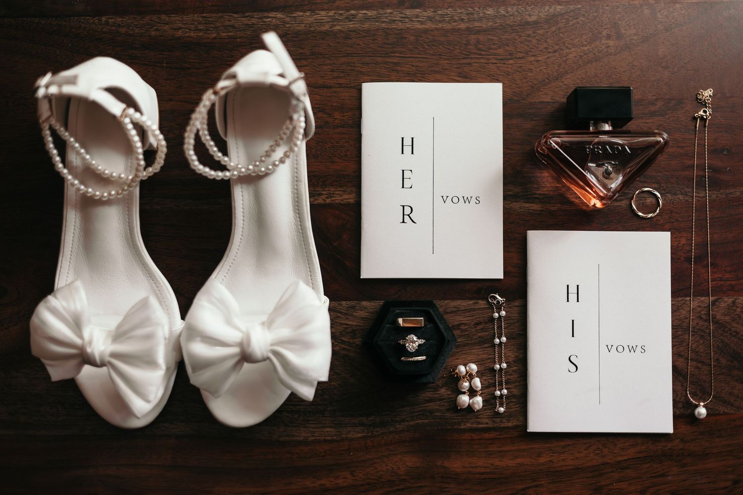 White bow heeled wedding shoes with pearl straps beside vow books, perfume, rings, and pearl jewelry on wood.