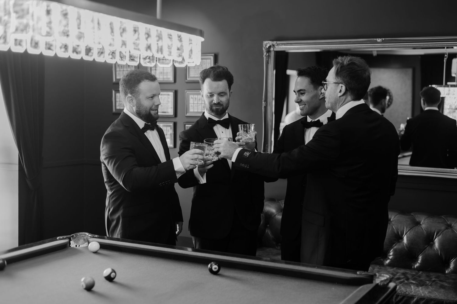 Groomsmen in tuxedos toasting with champagne glasses near a pool table in a stylish black and white wedding photo.