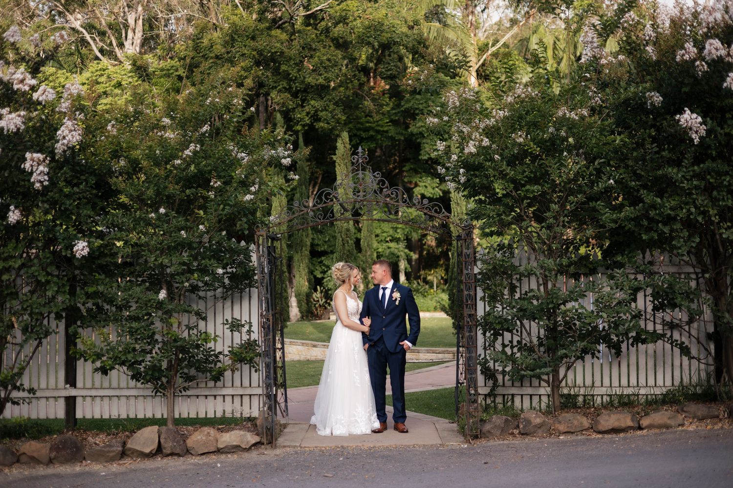 Bride and groom standing together at ornate iron garden gates surrounded by lush green trees and white flowering vines.
