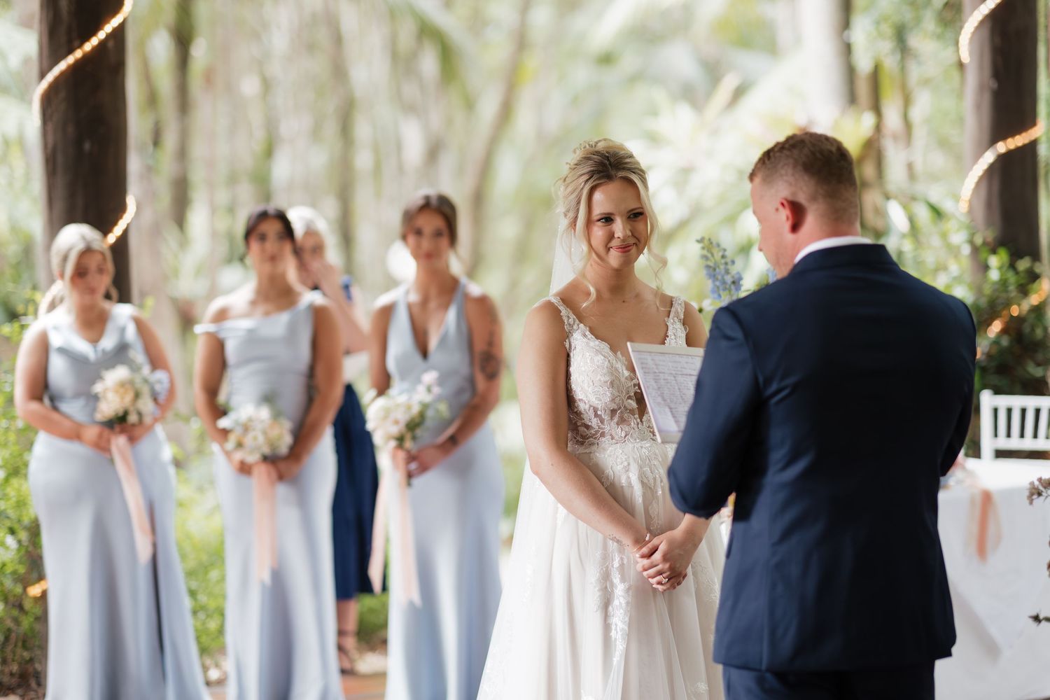 Bride smiling at groom during outdoor wedding ceremony with bridesmaids in blue dresses standing behind her.