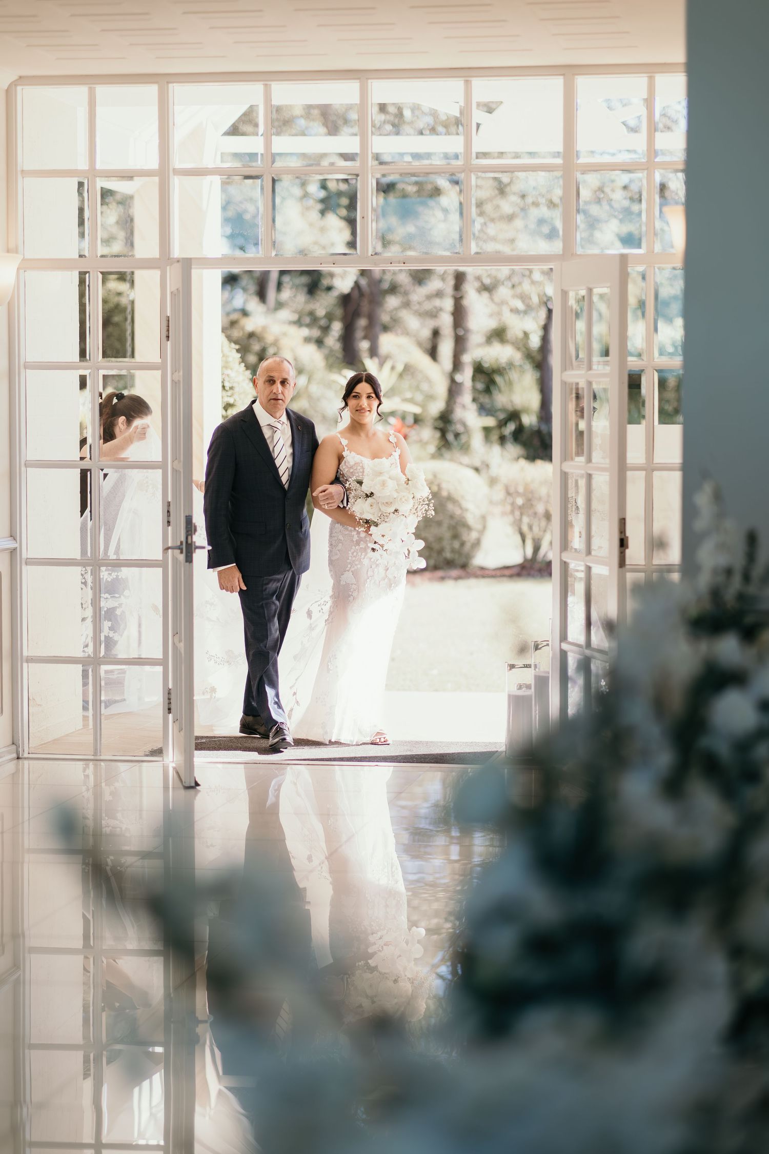 Elegant wedding couple enters sunlit venue through French doors in romantic garden setting.