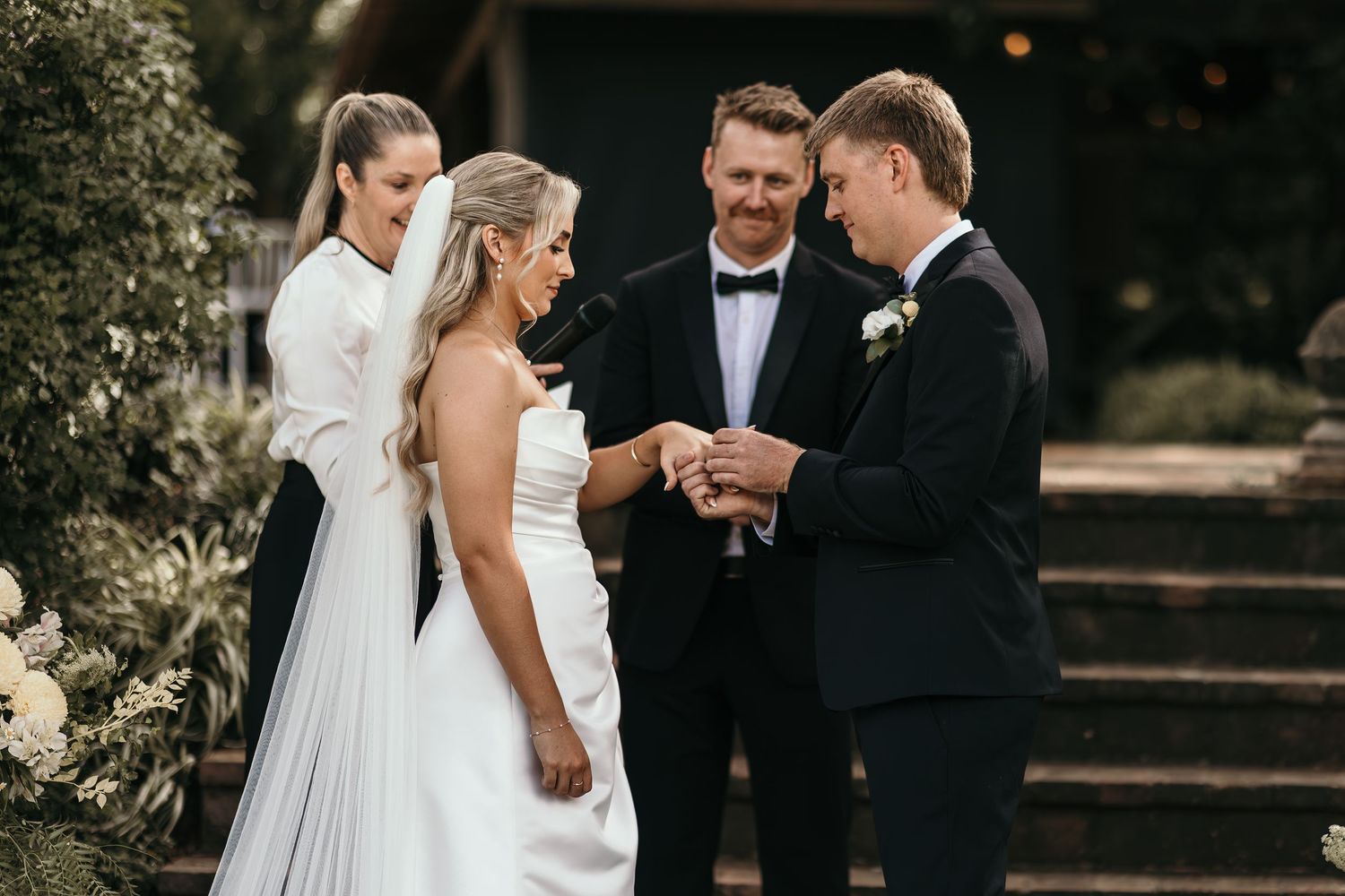 Groom placing wedding ring on bride's finger during outdoor ceremony with officiant and guest in background.