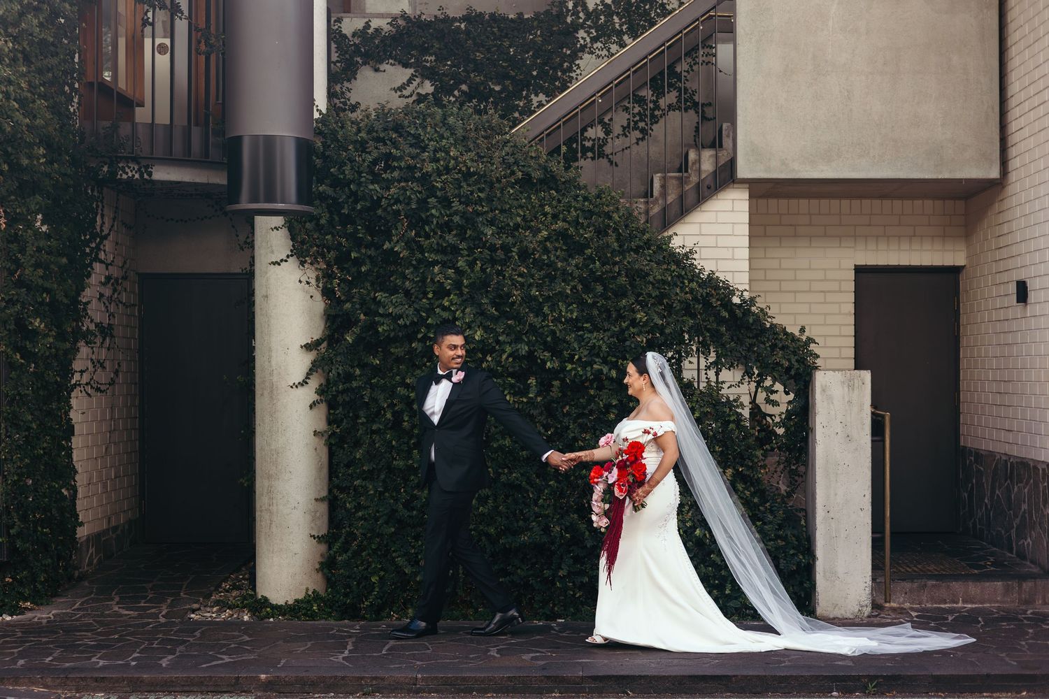 A bride in a white gown and groom in a tuxedo hold hands outside a building covered in lush green ivy.