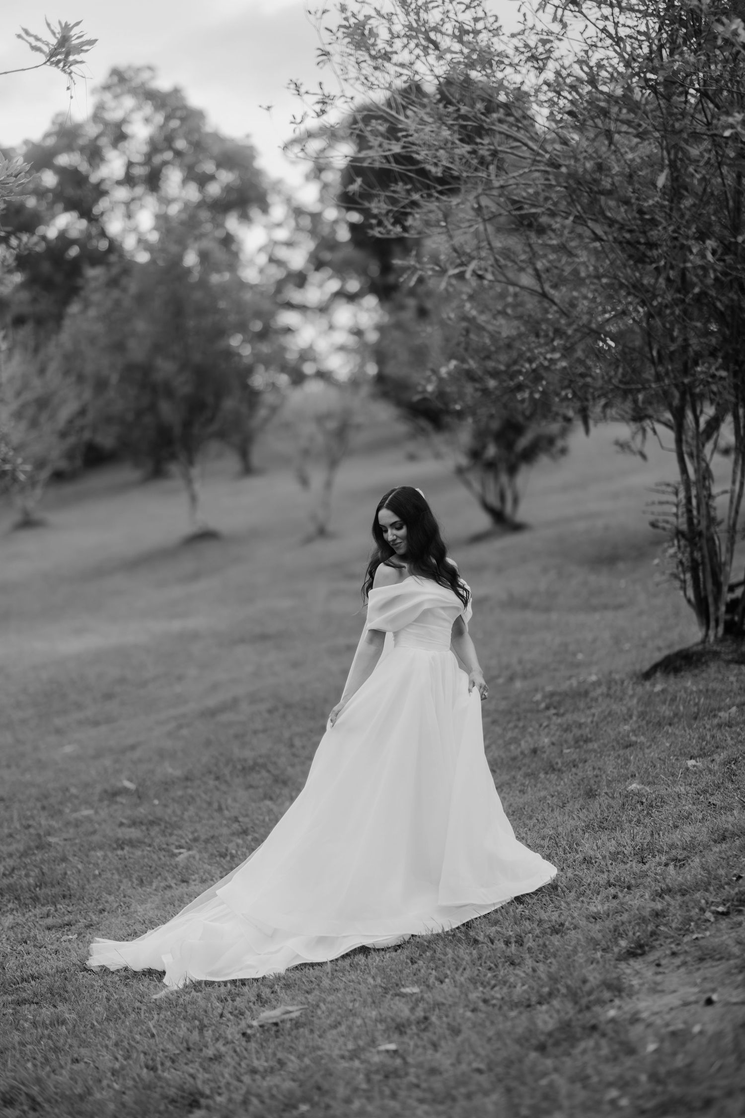 Black and white photo of a bride in an off-shoulder gown standing gracefully among trees in a lush garden.