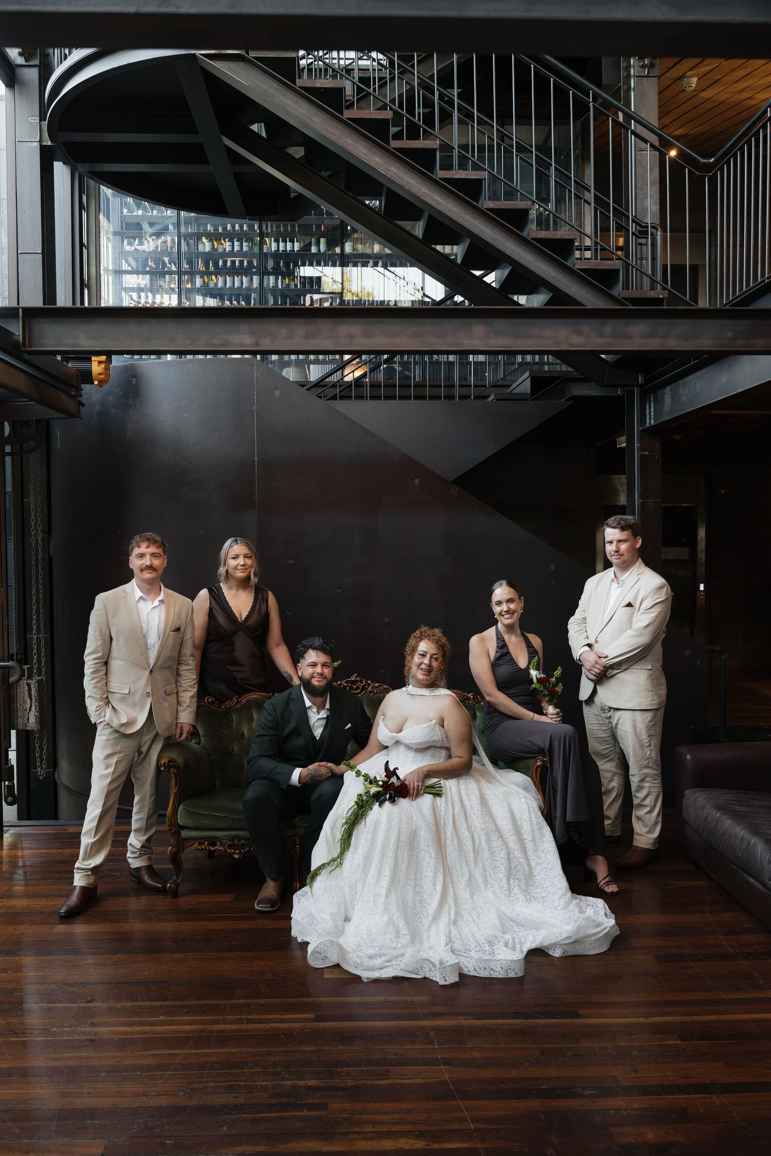 Wedding party poses elegantly in an industrial venue with dark walls and metal staircase, bride in white gown centered.