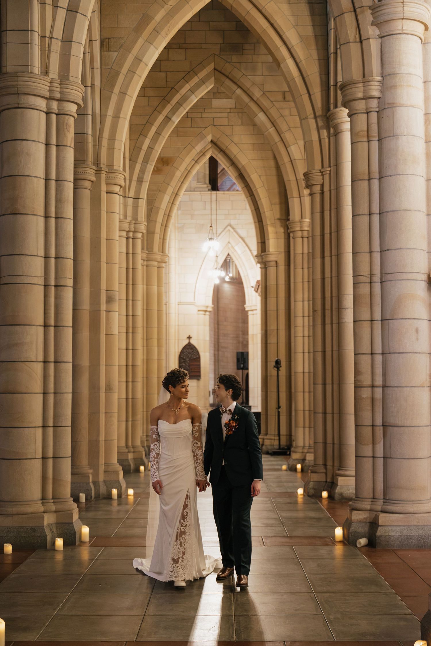 Two brides walk hand-in-hand through a candlelit Gothic cathedral aisle, one in a white lace gown and one in a black suit.