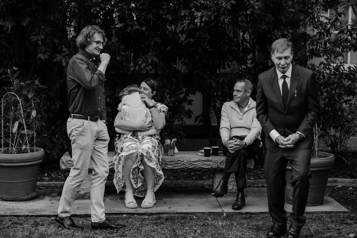Black and white outdoor photo of four people at a garden gathering, one man in a suit smiling on the right.