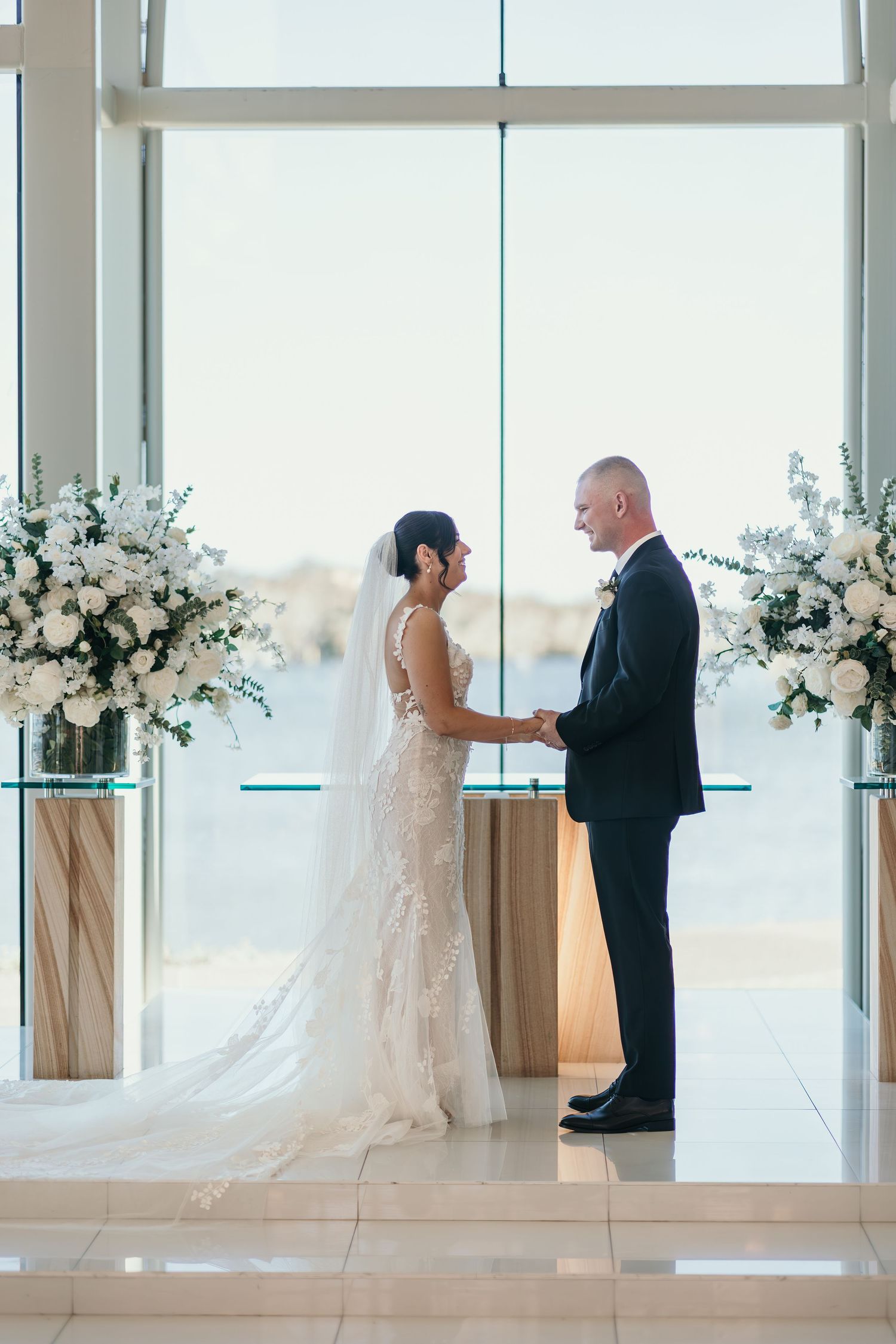 Elegant wedding ceremony in a bright white chapel with floral arrangements and floor-to-ceiling windows overlooking the ocean.
