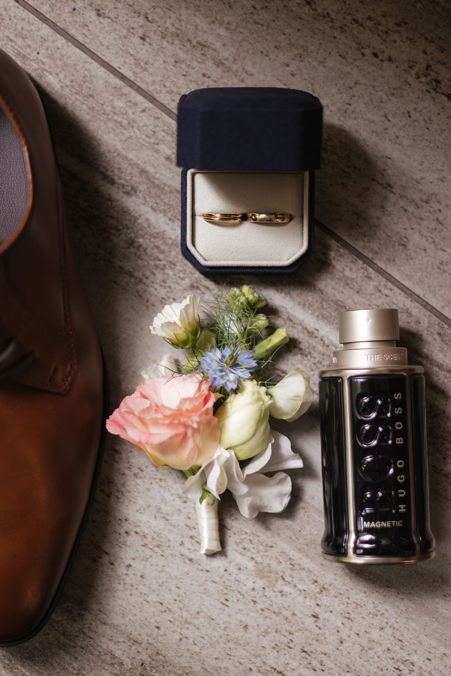 Wedding flat lay with gold rings in navy box, boutonniere, cologne bottle, and brown leather dress shoes.