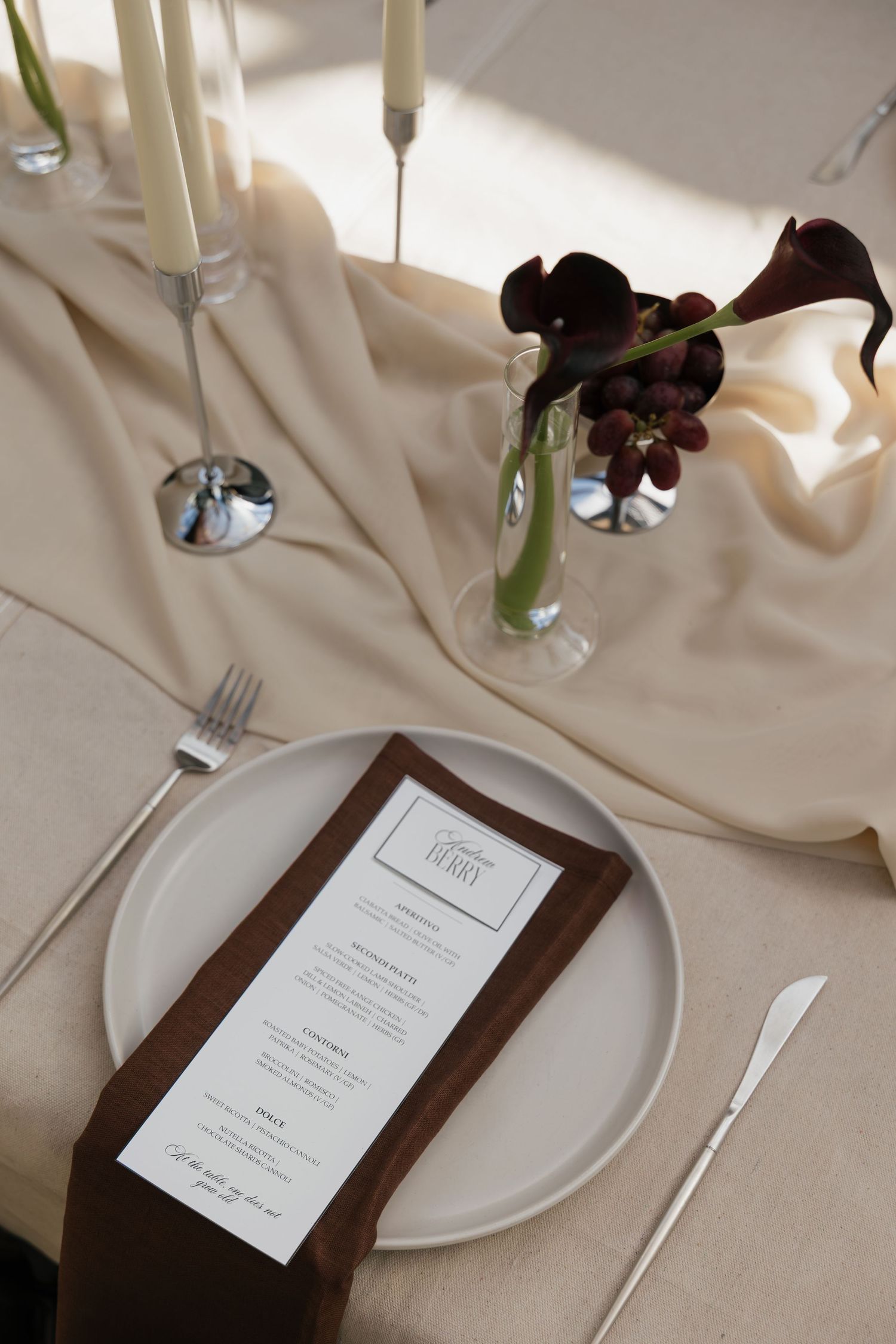 Elegant place setting with white plate, brown menu card, silver cutlery, candles, and dark floral arrangement on beige linen.