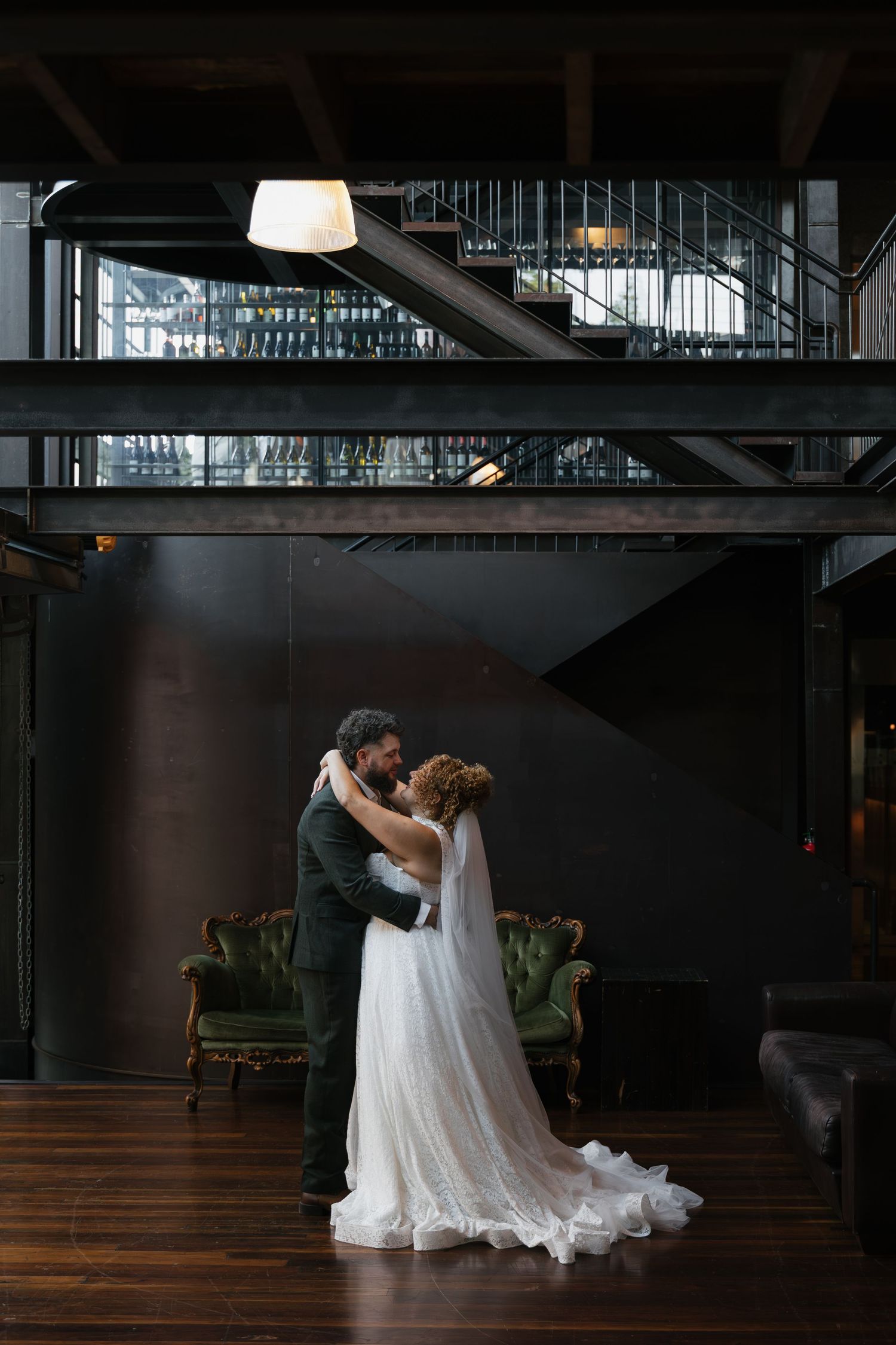 Bride and groom share first dance in elegant dark industrial venue with green velvet sofa and mezzanine staircase.