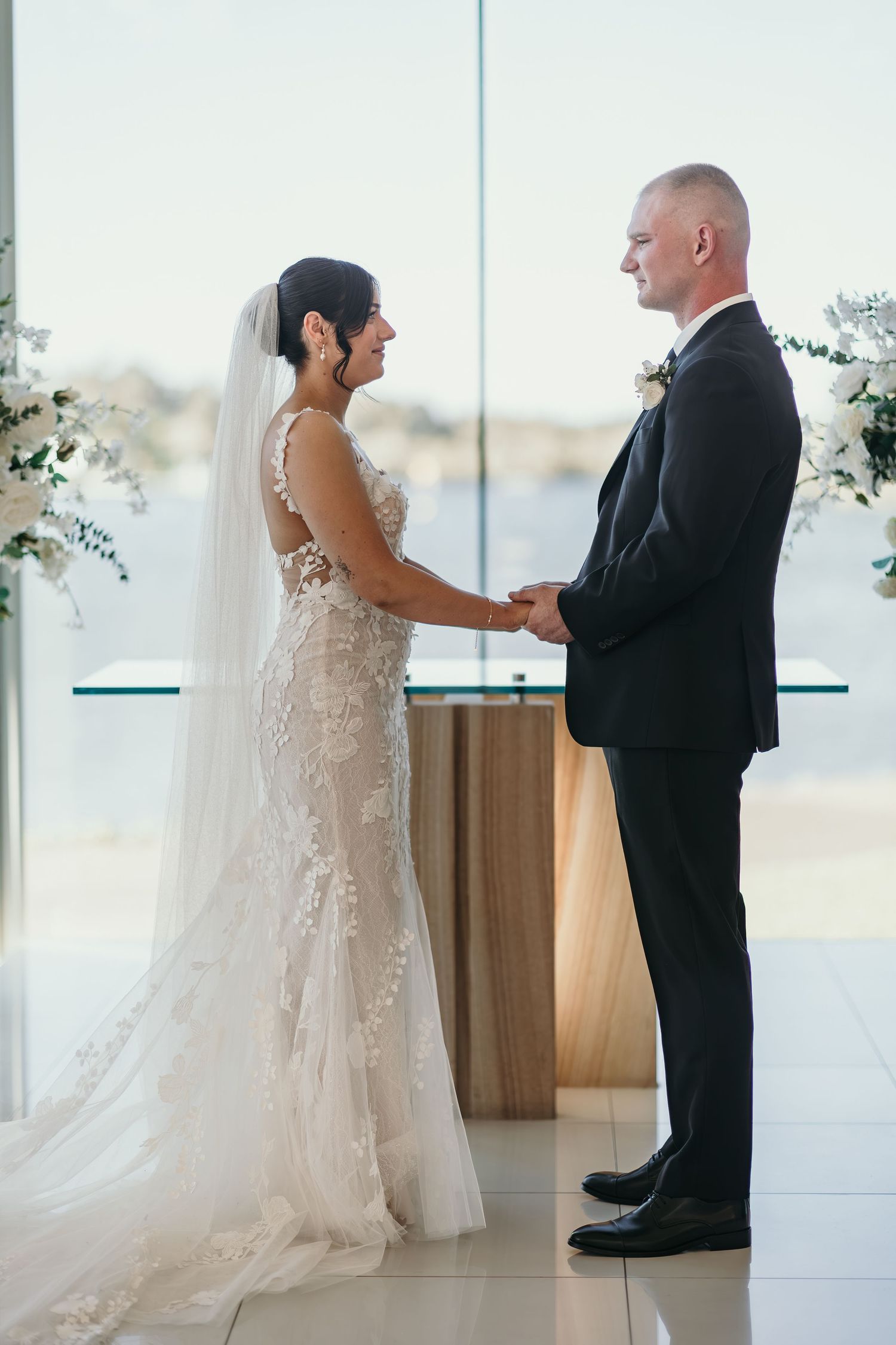 A couple shares an emotional first look moment in a bright modern venue with floor-to-ceiling windows and white floral decor.