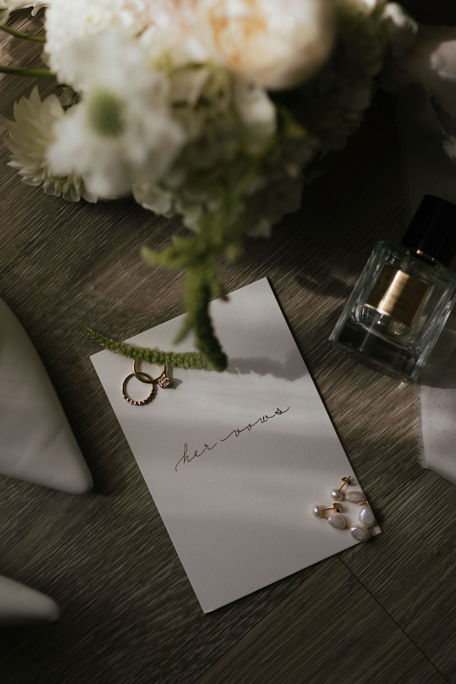 Elegant wedding flat lay with gold rings, pearl earrings, perfume bottle, and white floral bouquet on a wooden surface.