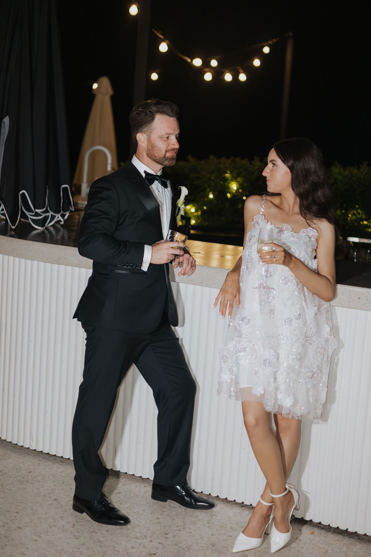 Elegant couple in formal attire sharing drinks at an outdoor evening wedding reception with string lights.