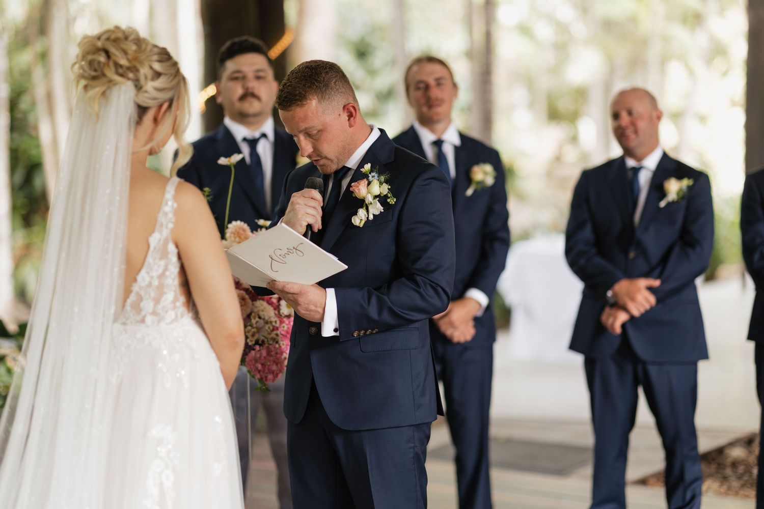 Groom reads wedding vows from paper while bride listens, groomsmen stand in background at outdoor ceremony.