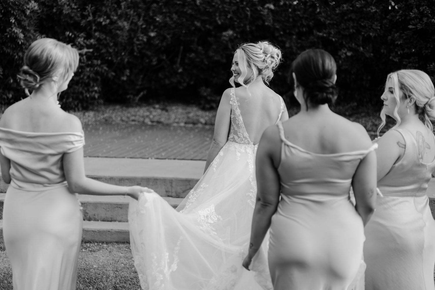 Black and white photo of a bride in lace gown with four bridesmaids in white dresses walking together outdoors.