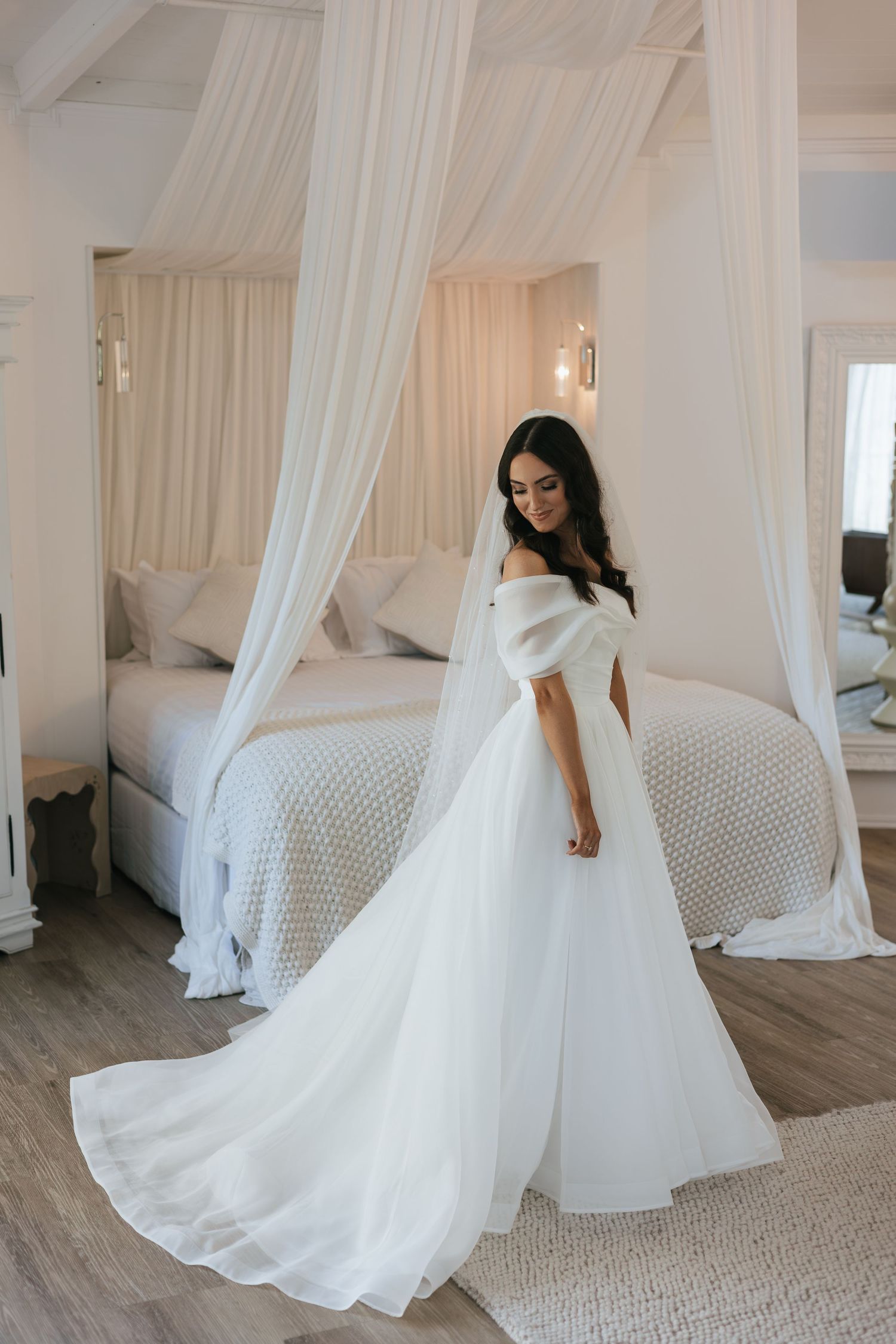 Bride in elegant off-shoulder white ball gown with long train stands in a luxurious canopy bedroom.