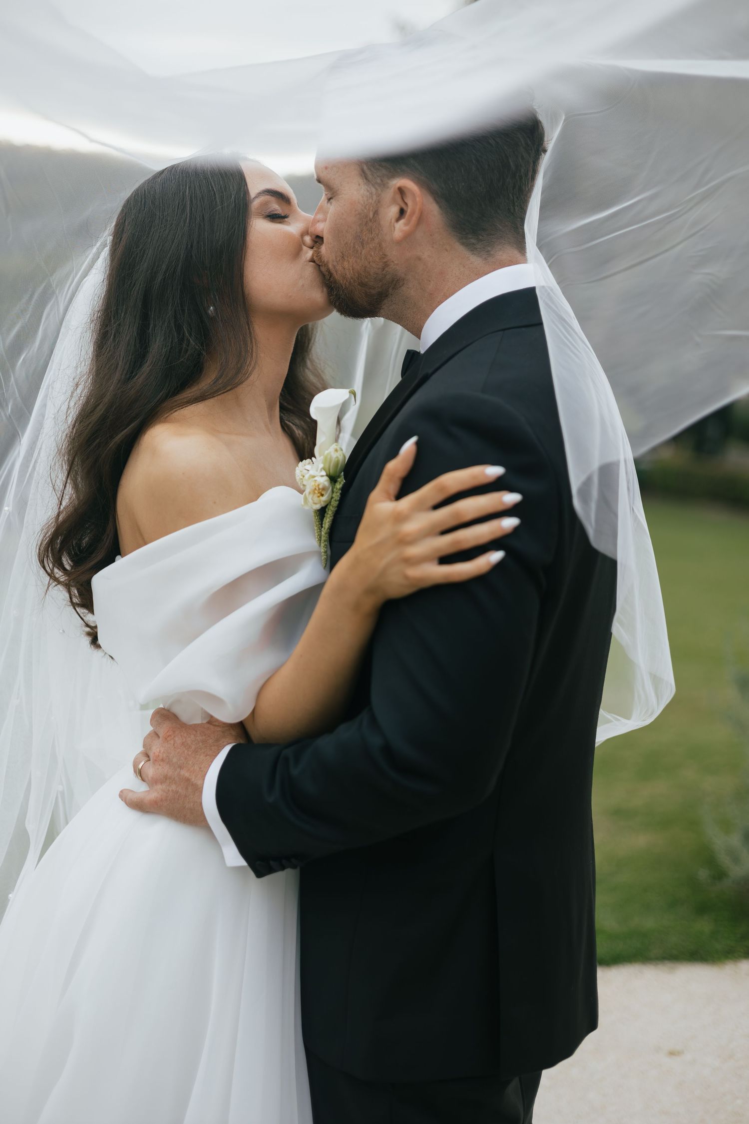 Bride and groom sharing a romantic kiss under a flowing white veil on their wedding day outdoors.