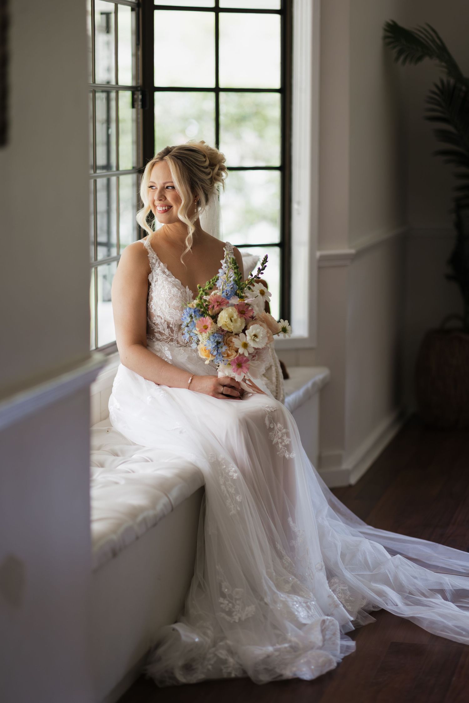Smiling bride in lace wedding gown holding colorful bouquet, seated by window in elegant indoor setting.