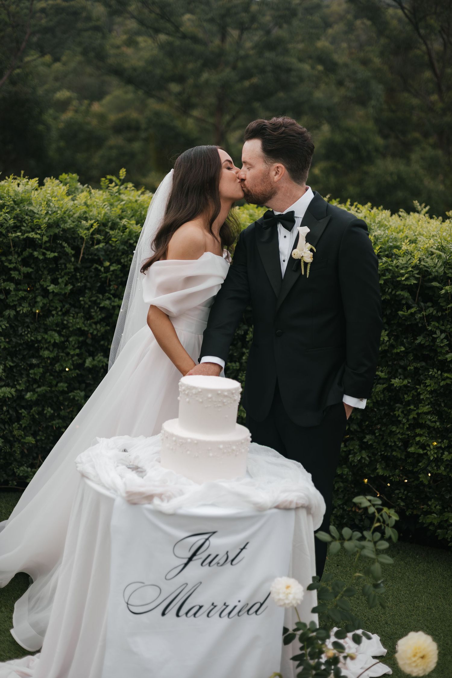 Newlyweds kissing behind a white tiered wedding cake on a Just Married decorated table in a garden setting.